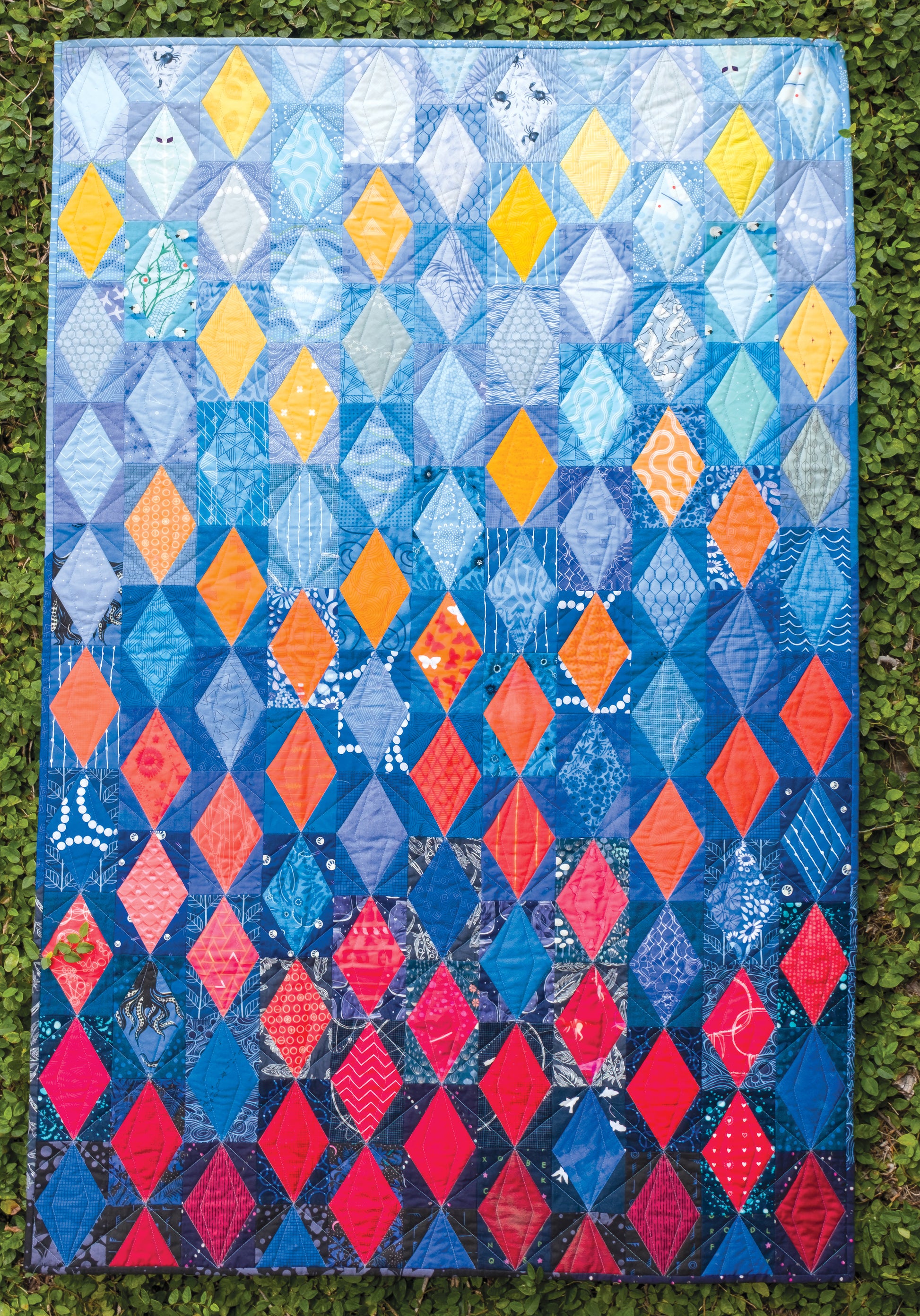 Experience the vibrant design of the *Using Scraps in Modern Quilts: Design Lab* by Creative Spark—a lively quilt showcasing diamond patterns in blue, yellow, orange, and red, artfully displayed on lush green grass. The inventive use of negative space enhances its modern aesthetic and brings each color to life.