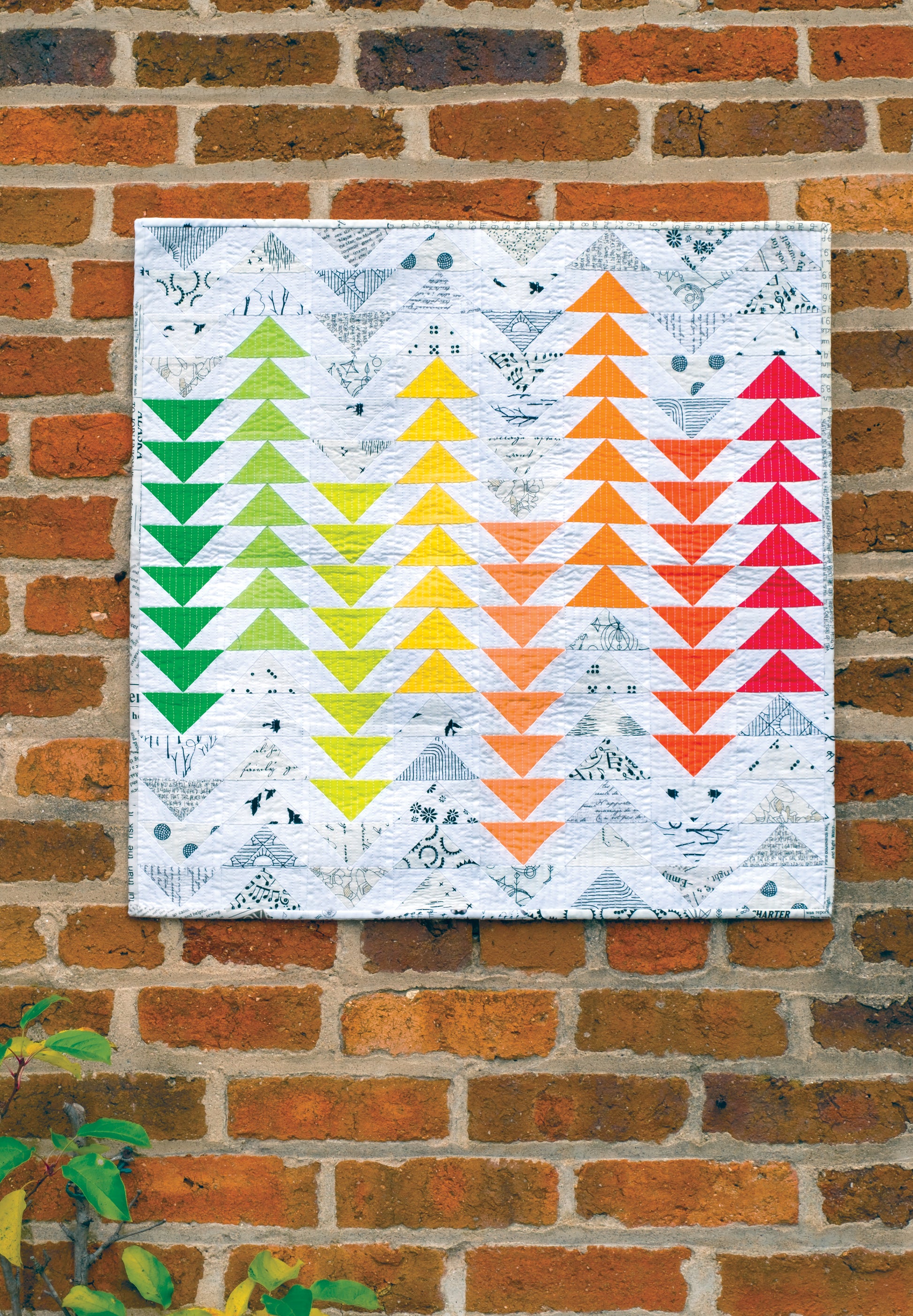 Showcasing skillful use of negative space, the "Using Scraps in Modern Quilts: Design Lab" by Creative Spark features a modern quilt with arrow-like patterns in green, yellow, orange, and red arranged in columns. The quilt is displayed against a brick wall with some leaves visible at the bottom left corner.