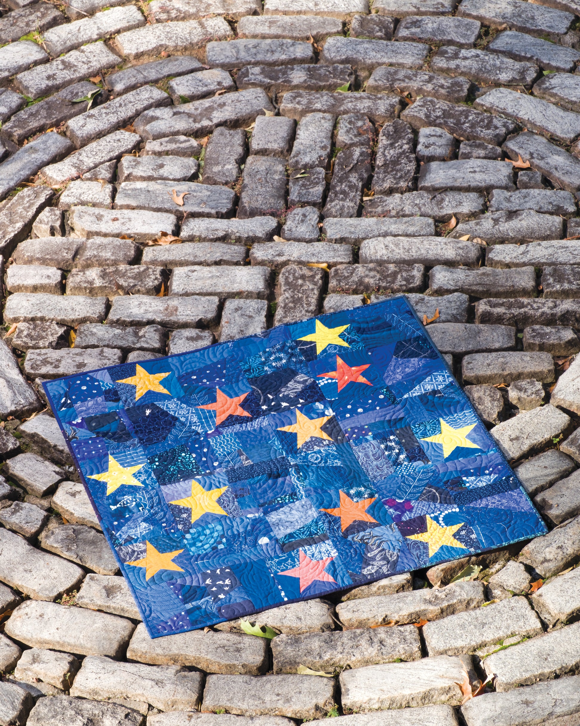 A modern quilt, designed using techniques from Creative Spark’s "Using Scraps in Modern Quilts: Design Lab", features blue fabric accented with yellow and orange star motifs, ghost blocks, and inventive negative space; it is displayed on a circular stone path with a spiral pattern.