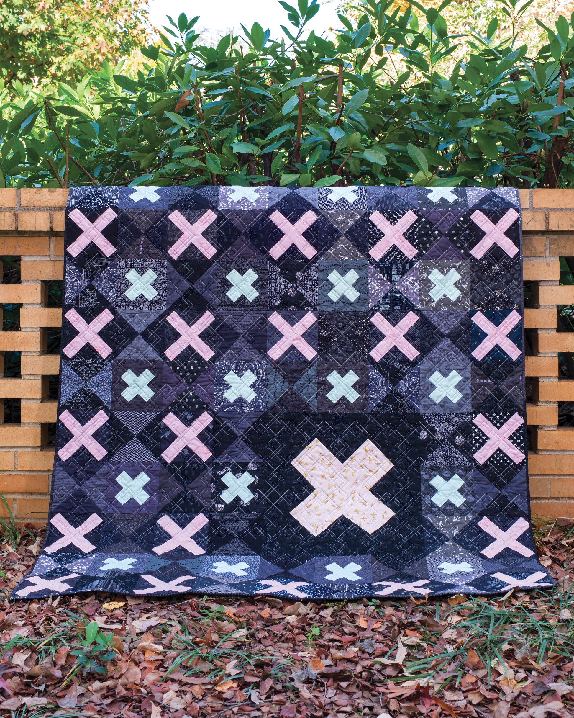 A quilt from Creative Spark's "Using Scraps in Modern Quilts: Design Lab" is draped over a brick wall, showcasing X-shaped patterns reminiscent of ghost blocks in pastel colors on a dark background. Green shrubs and fallen leaves are visible in the background, highlighting how this modern quilt skillfully uses negative space to make its design stand out beautifully.