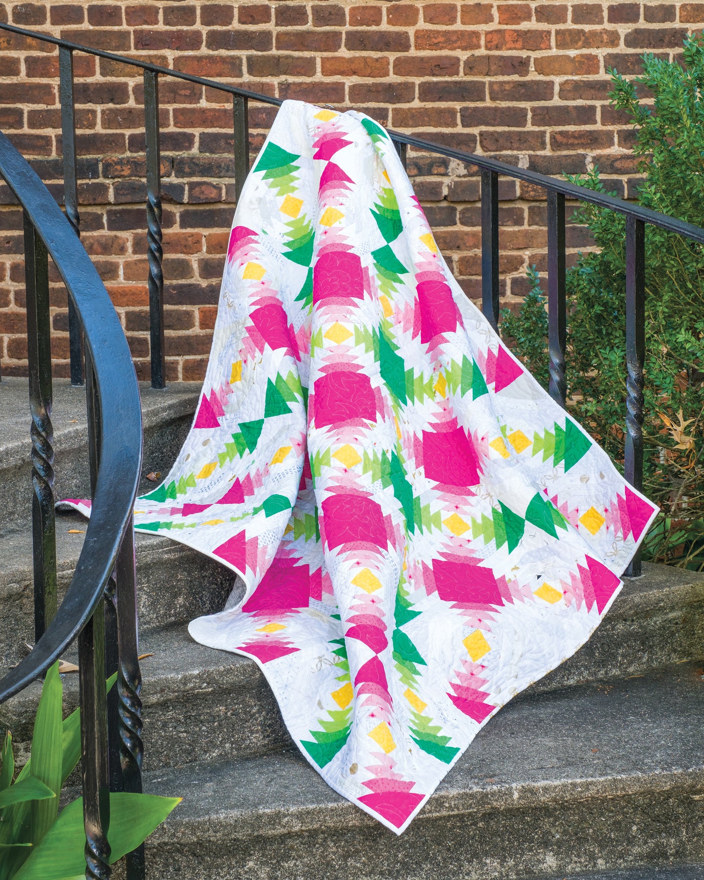 A vibrant quilt, designed with pink, yellow, green, and white geometric patterns and incorporating skillful ghost blocks for intriguing negative space, drapes over a black iron railing on stone steps outdoors with a brick wall and greenery in the background. This fresh take on modern quilting is featured in "Using Scraps in Modern Quilts: Design Lab" by Creative Spark.