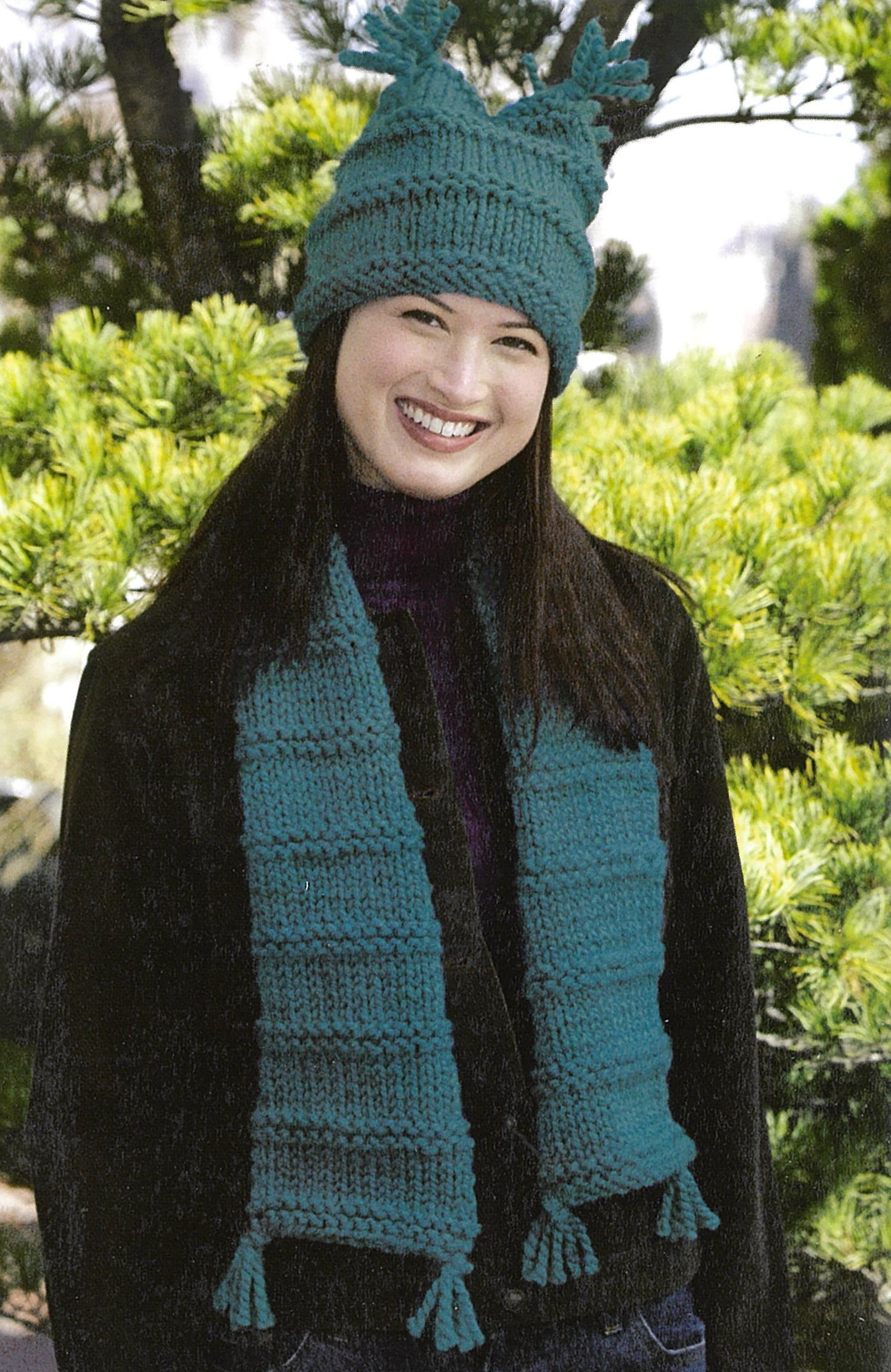 A smiling person stands outdoors in front of green foliage, wearing a teal knit hat with tassels and a matching scarf made using patterns from Learn To Knit—Lion Brand Yarn Booklet by Leisure Arts.