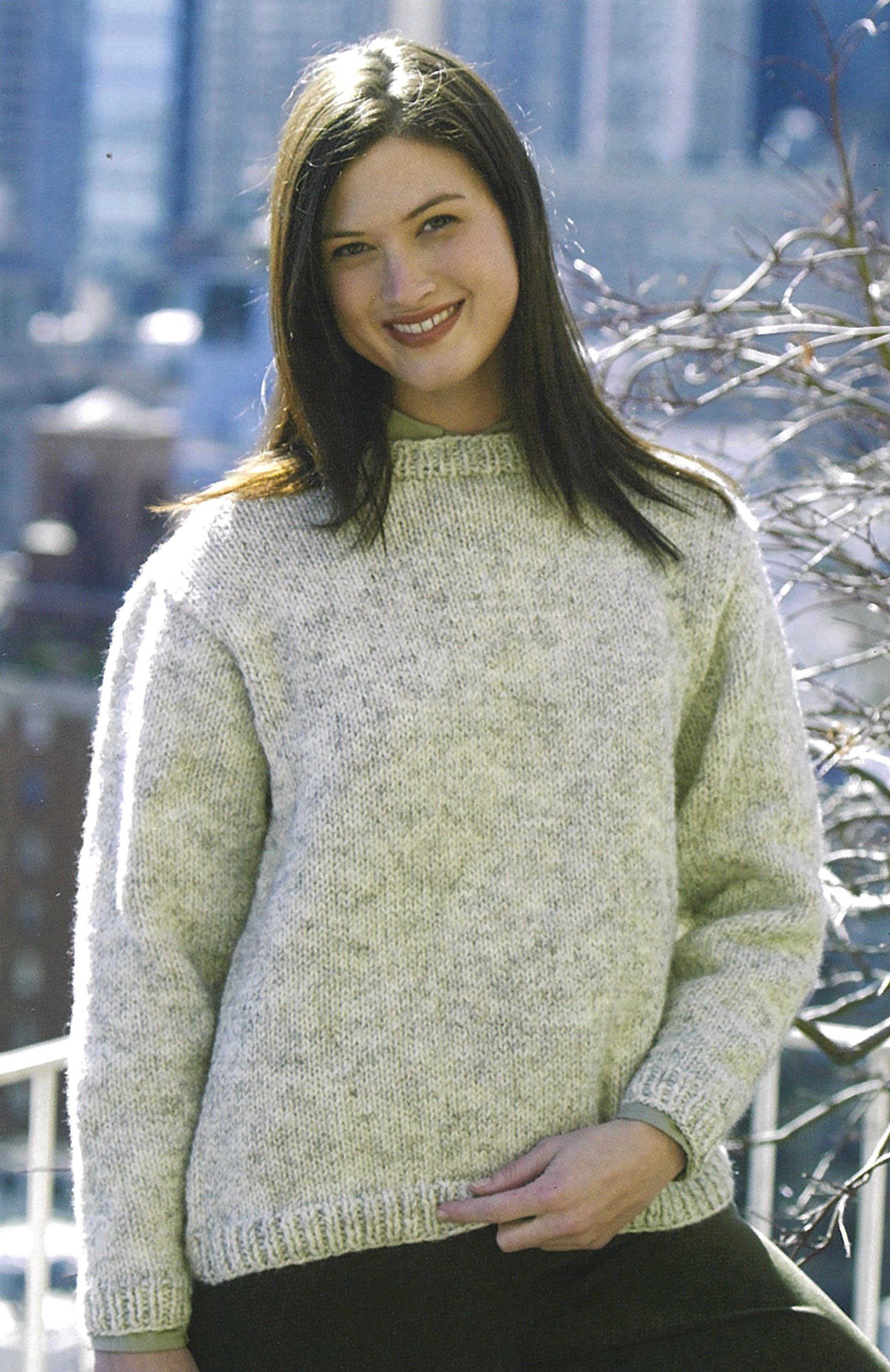 A woman with long brown hair smiles outdoors in a light gray sweater, perfect for beginners using the "Learn To Knit—Lion Brand Yarn Booklet" from Leisure Arts, with a blurred cityscape in the background.
