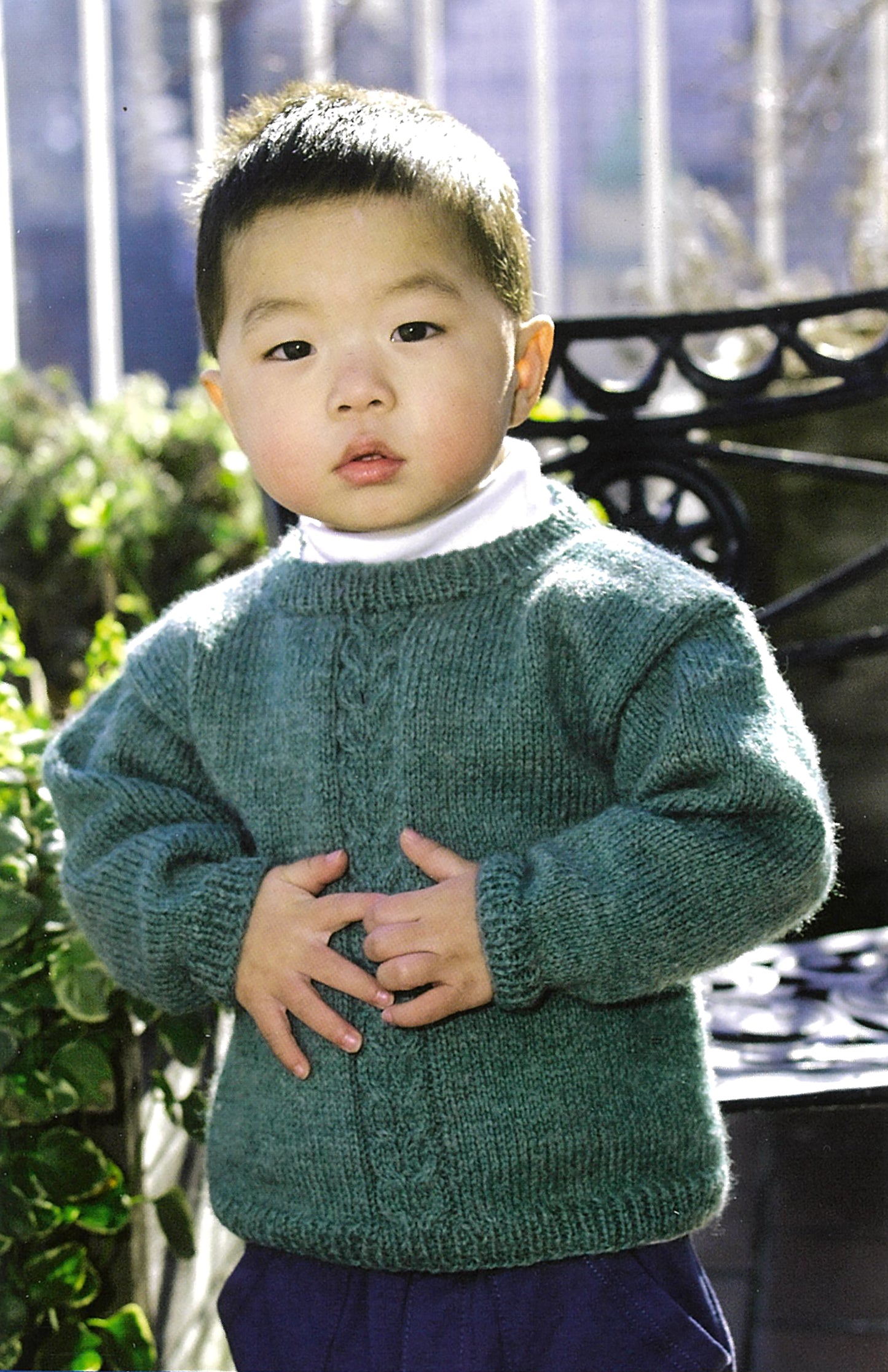 A young child with short hair stands outdoors, wearing a green sweater that could be made from patterns in the "Learn To Knit—Lion Brand Yarn Booklet" by Leisure Arts, with greenery and a metal bench in the background.