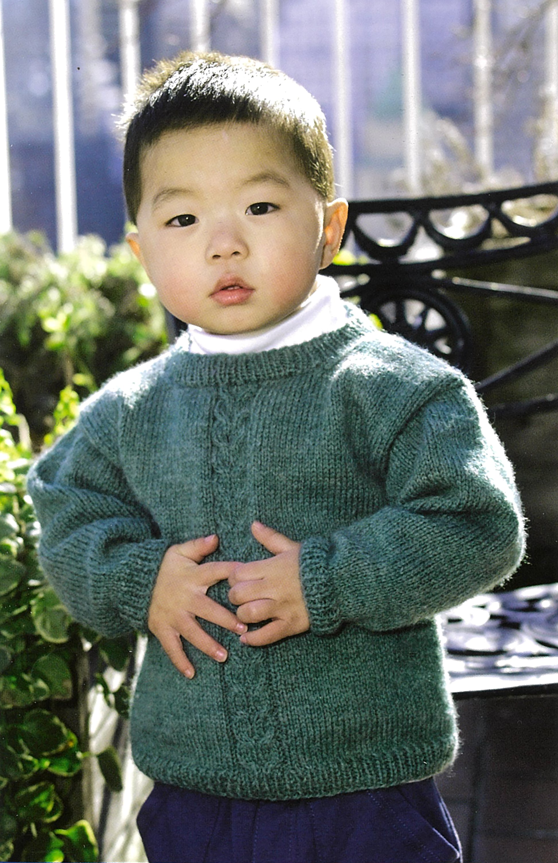 A young child with short hair stands outdoors, wearing a green sweater that could be made from patterns in the "Learn To Knit—Lion Brand Yarn Booklet" by Leisure Arts, with greenery and a metal bench in the background.