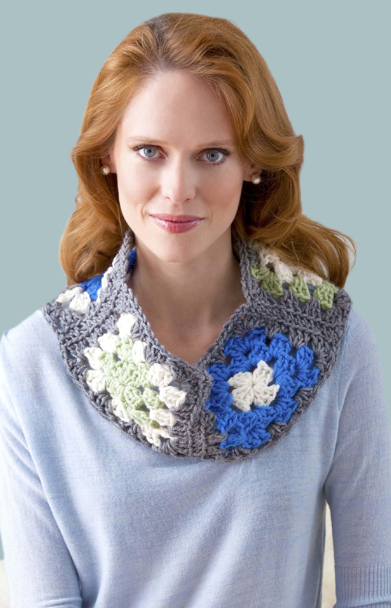 A red-haired woman in a light blue sweater wears a crocheted cowl made with the "You Can Do It Granny Squares" pattern by Leisure Arts, featuring blue, white, green, and gray granny squares against a light blue background.