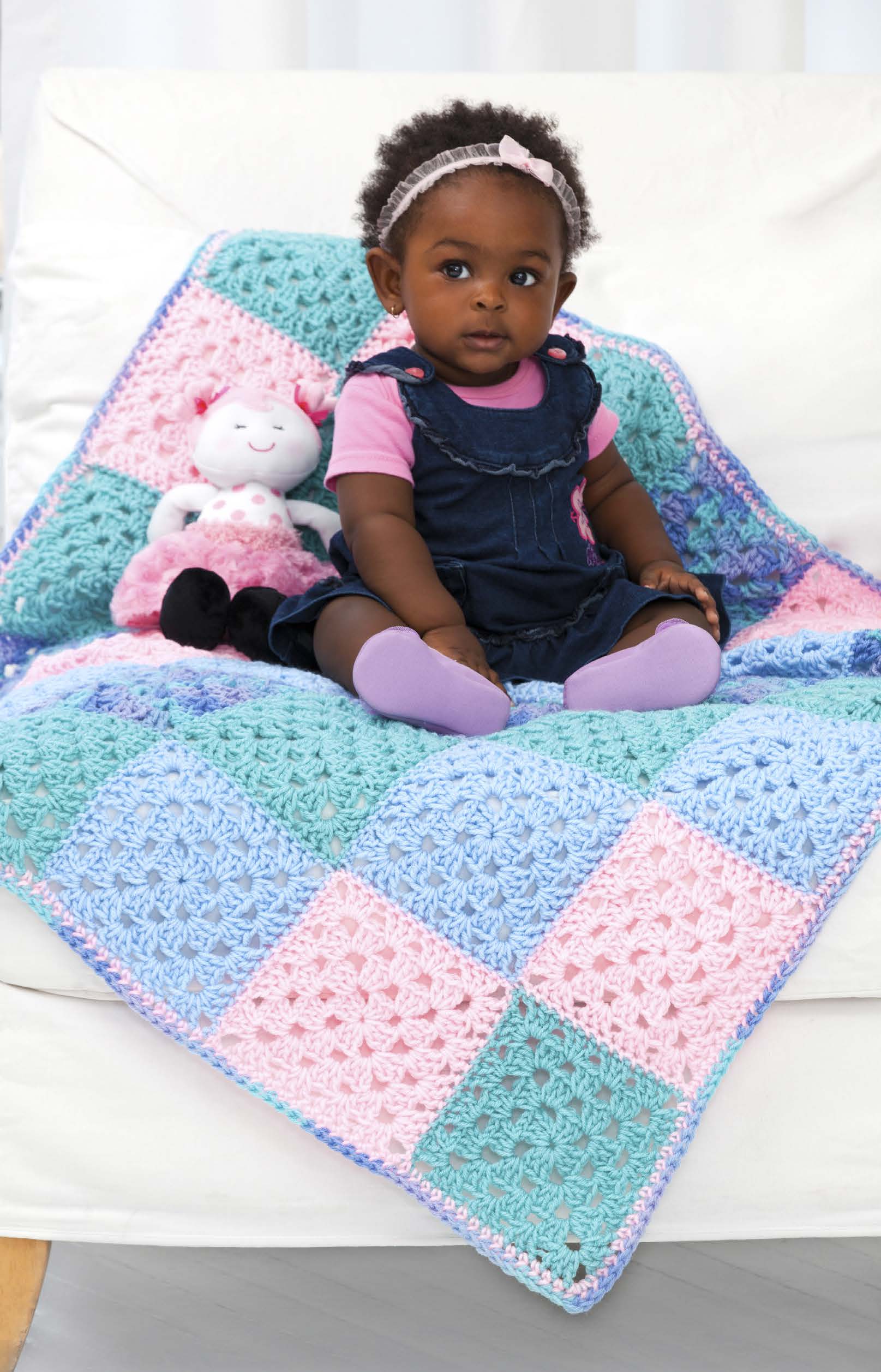 A baby sits on a white couch, wearing a pink shirt, denim jumper, lavender socks, and a pink headband. She’s on a pastel crocheted blanket inspired by Leisure Arts’ You Can Do It Granny Squares—perfect for beginner crochet projects—beside a stuffed cat.