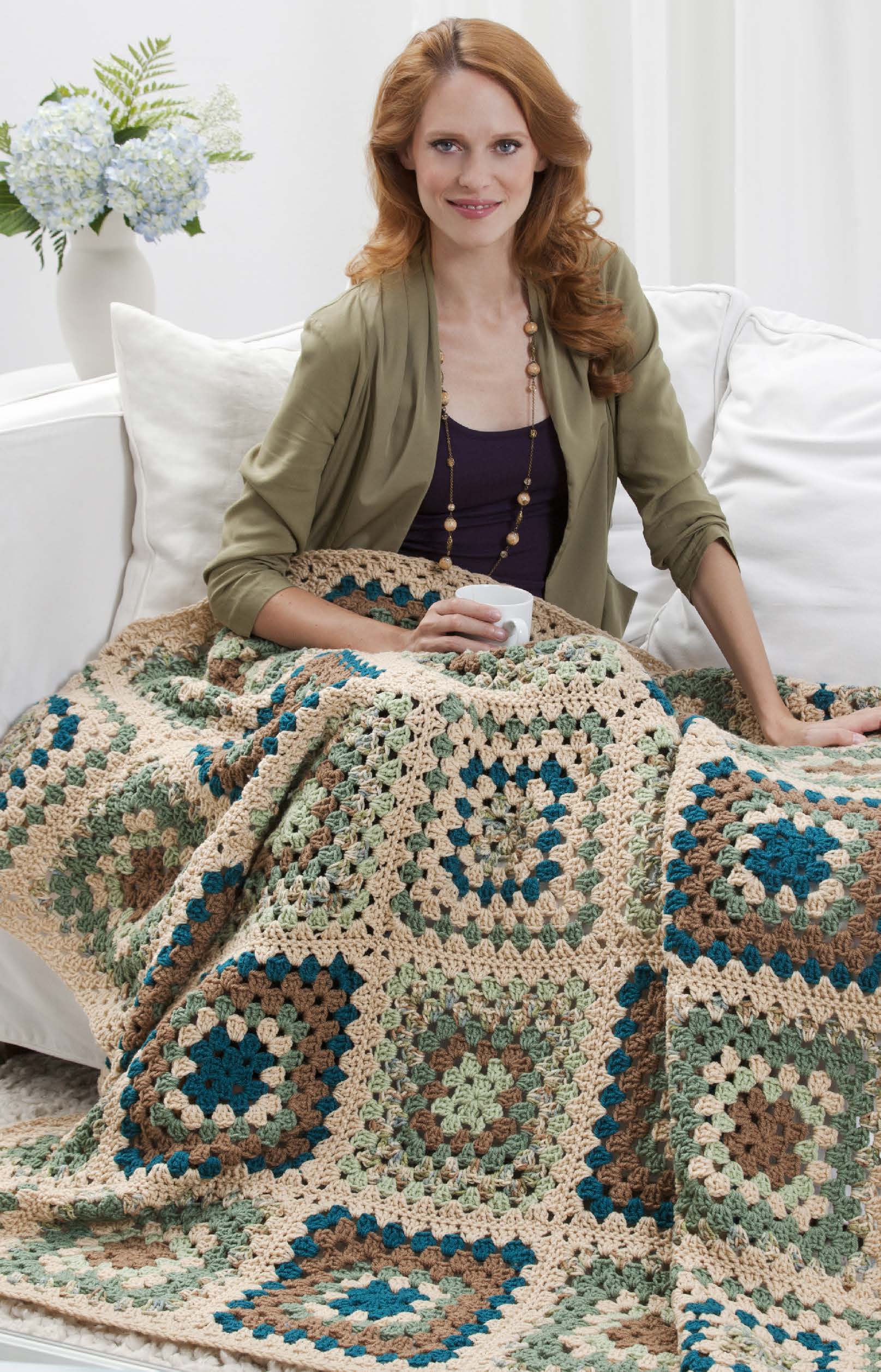 A woman with long red hair sits on a white couch, smiling and holding a mug. She is wrapped in a large, multicolored blanket made from the "You Can Do It Granny Squares" kit by Leisure Arts, featuring blue, green, beige, and tan granny squares.