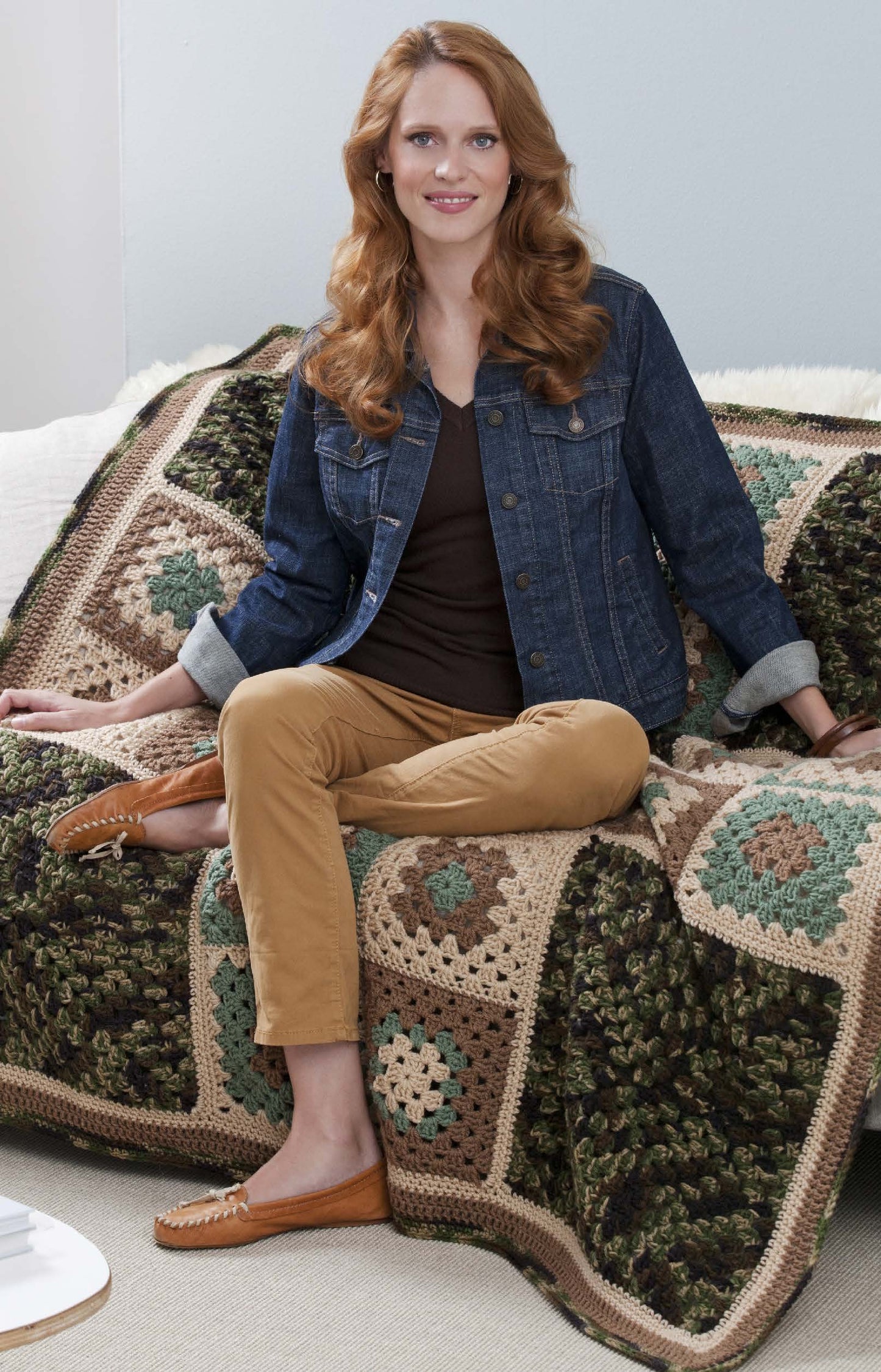 A woman with long red hair, in a denim jacket and tan pants, sits on a couch with a cream and green crochet blanket made from Leisure Arts’ You Can Do It Granny Squares, perfect for beginner crochet projects, smiling at the camera.