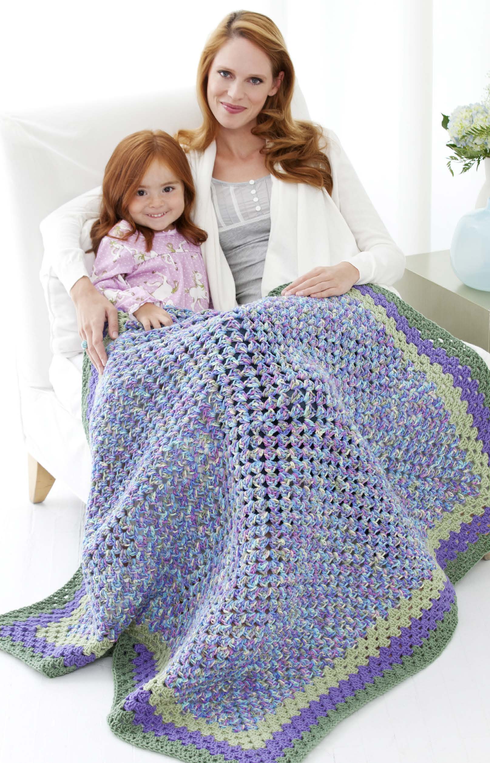 A woman and young girl smile on a white chair with the colorful "You Can Do It Granny Squares" crochet blanket by Leisure Arts in purple, blue, and green on their laps. A vase of flowers sits on the table beside them.