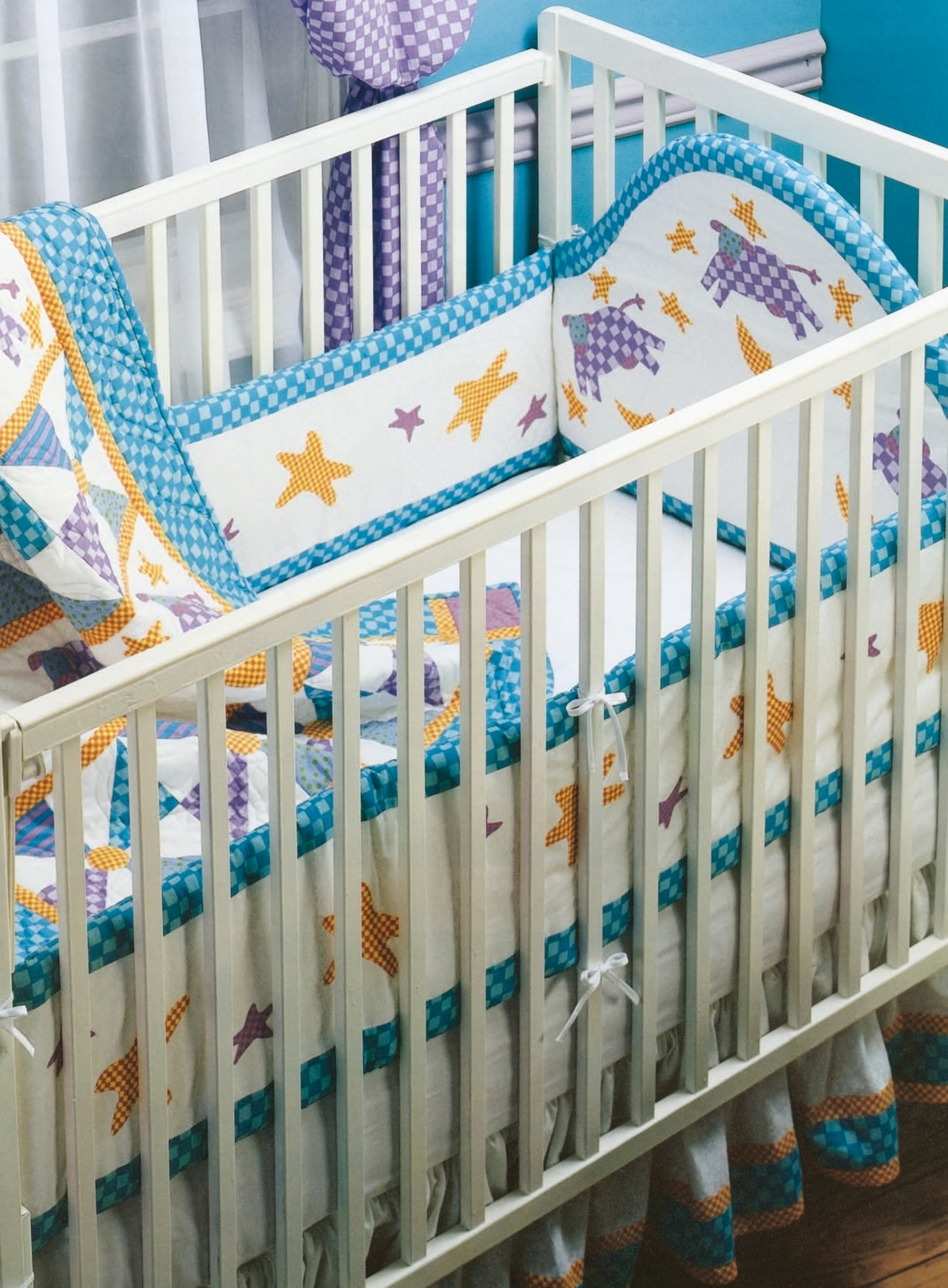 A white baby crib featuring a Rock-a-Bye Quilts bumper pad by Leisure Arts, decorated with yellow stars and purple patchwork, set against a blue wall with a window. The crib includes a white mattress and patterned skirt.