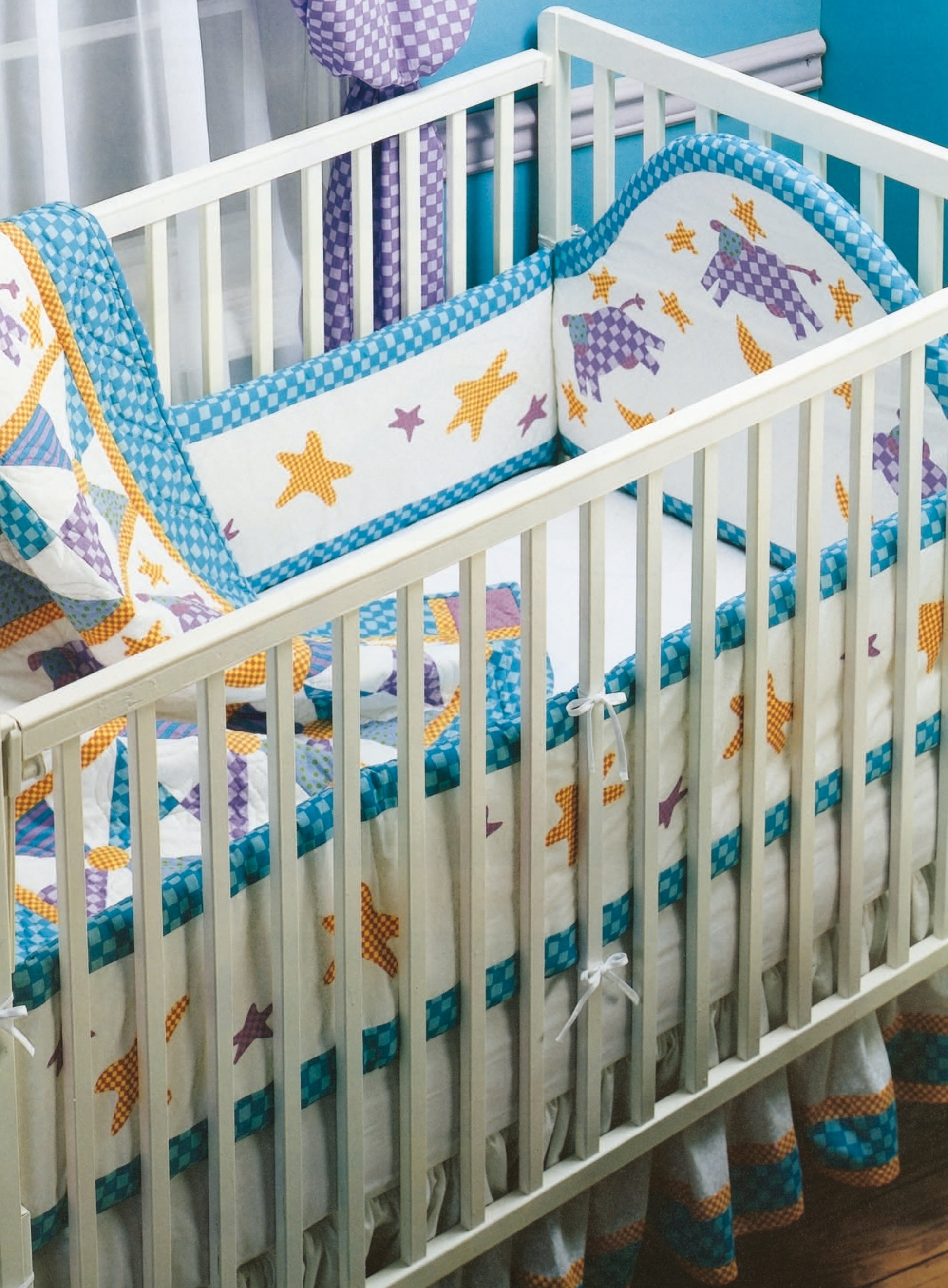 A white baby crib featuring a Rock-a-Bye Quilts bumper pad by Leisure Arts, decorated with yellow stars and purple patchwork, set against a blue wall with a window. The crib includes a white mattress and patterned skirt.