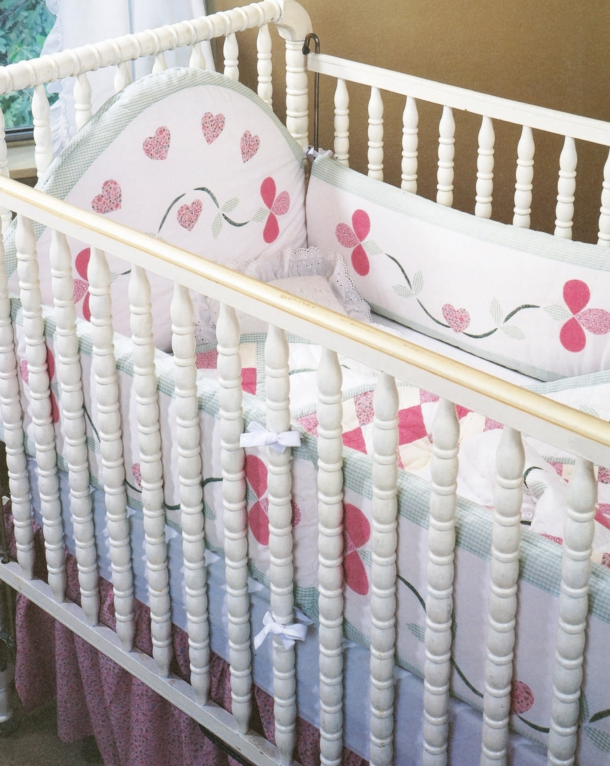 The Rock-a-Bye Quilts by Leisure Arts features a white wooden baby crib with spindled sides, a floral bumper, pink ruffled skirt, and bedding adorned with pink hearts and flowers. Sunlight streams through a nearby window.