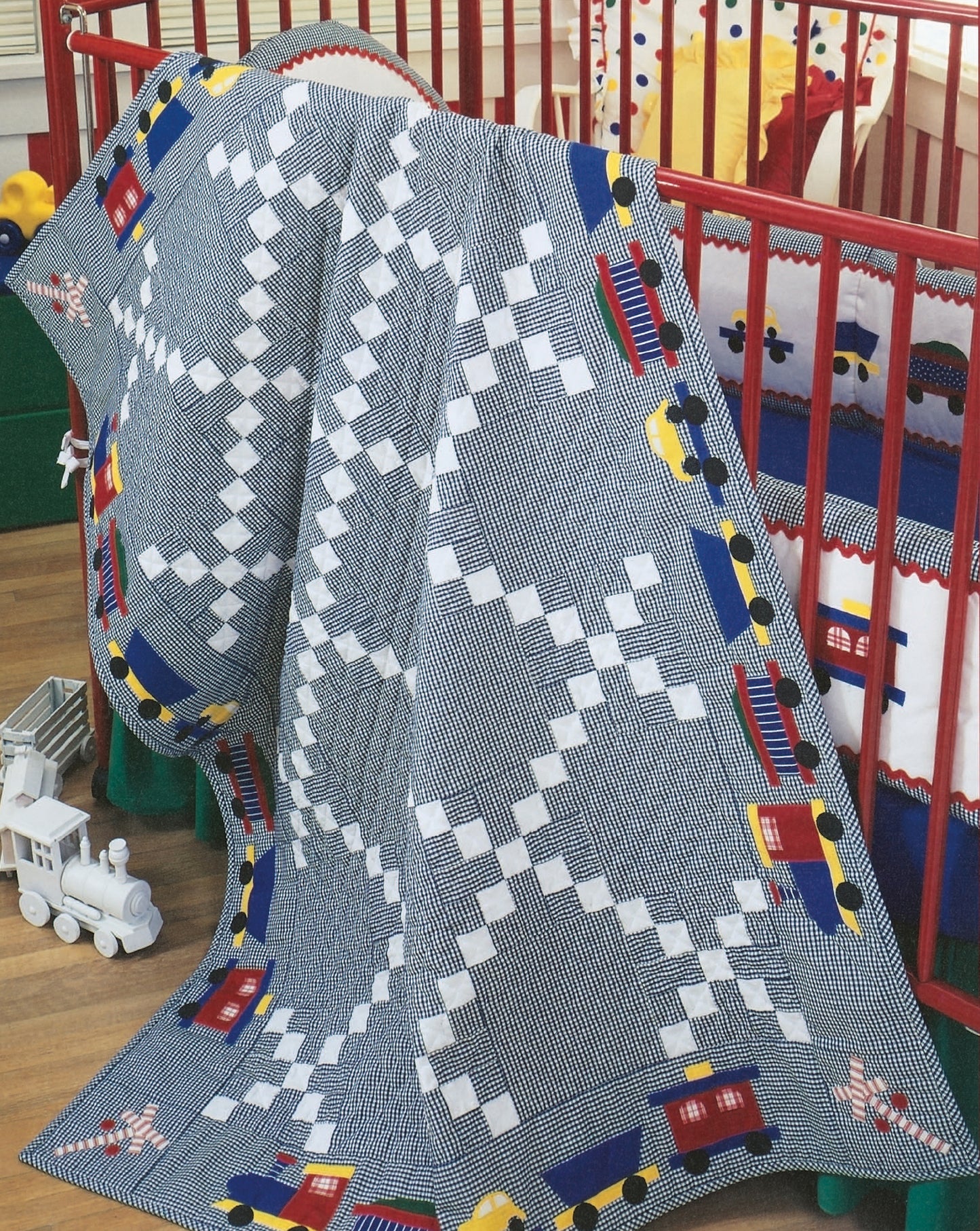 The Rock-a-Bye Quilts by Leisure Arts features a blue and white geometric baby quilt with colorful toy trains, draped over a red crib, with a white toy train resting on the wooden floor nearby.