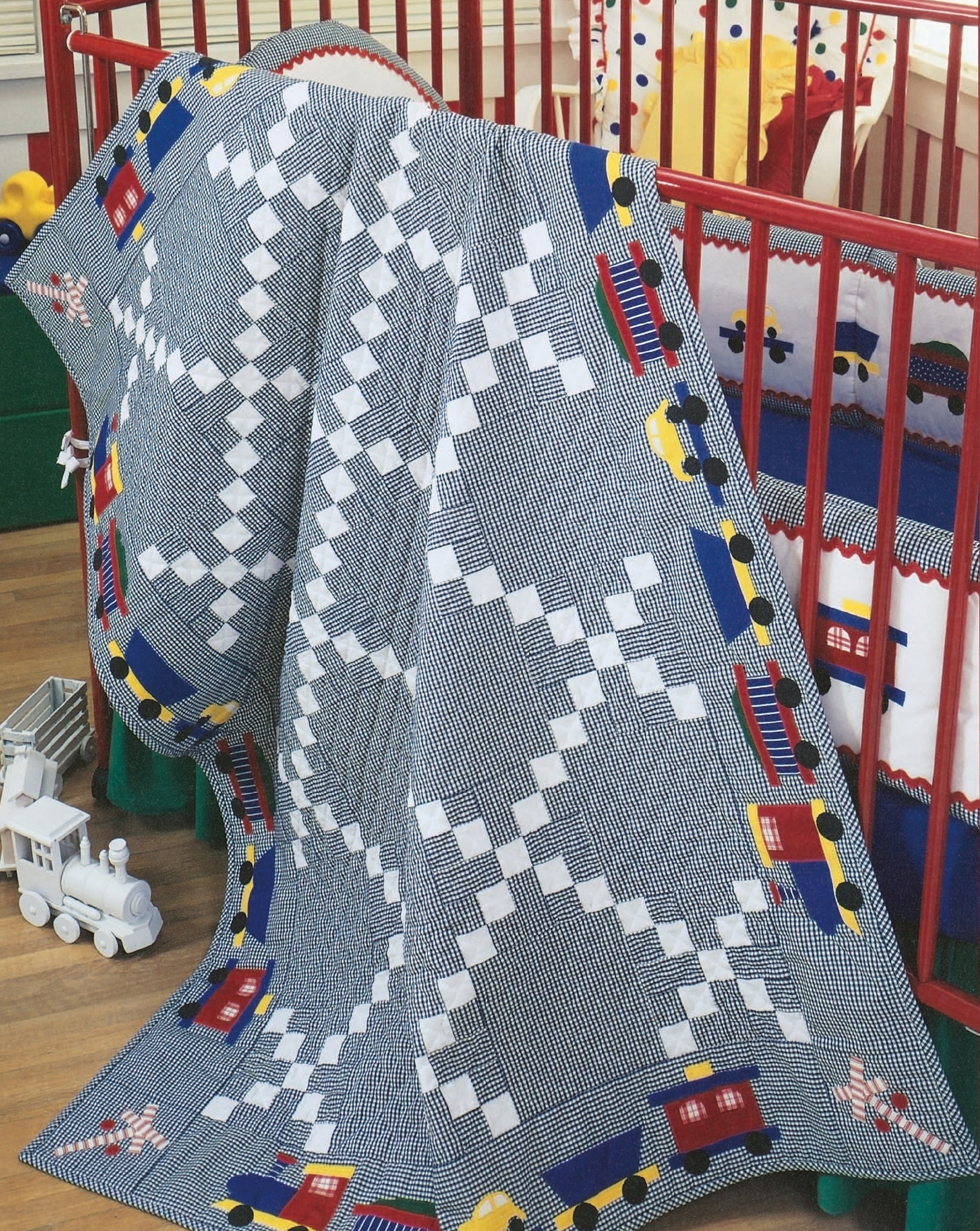 The Rock-a-Bye Quilts by Leisure Arts features a blue and white geometric baby quilt with colorful toy trains, draped over a red crib, with a white toy train resting on the wooden floor nearby.