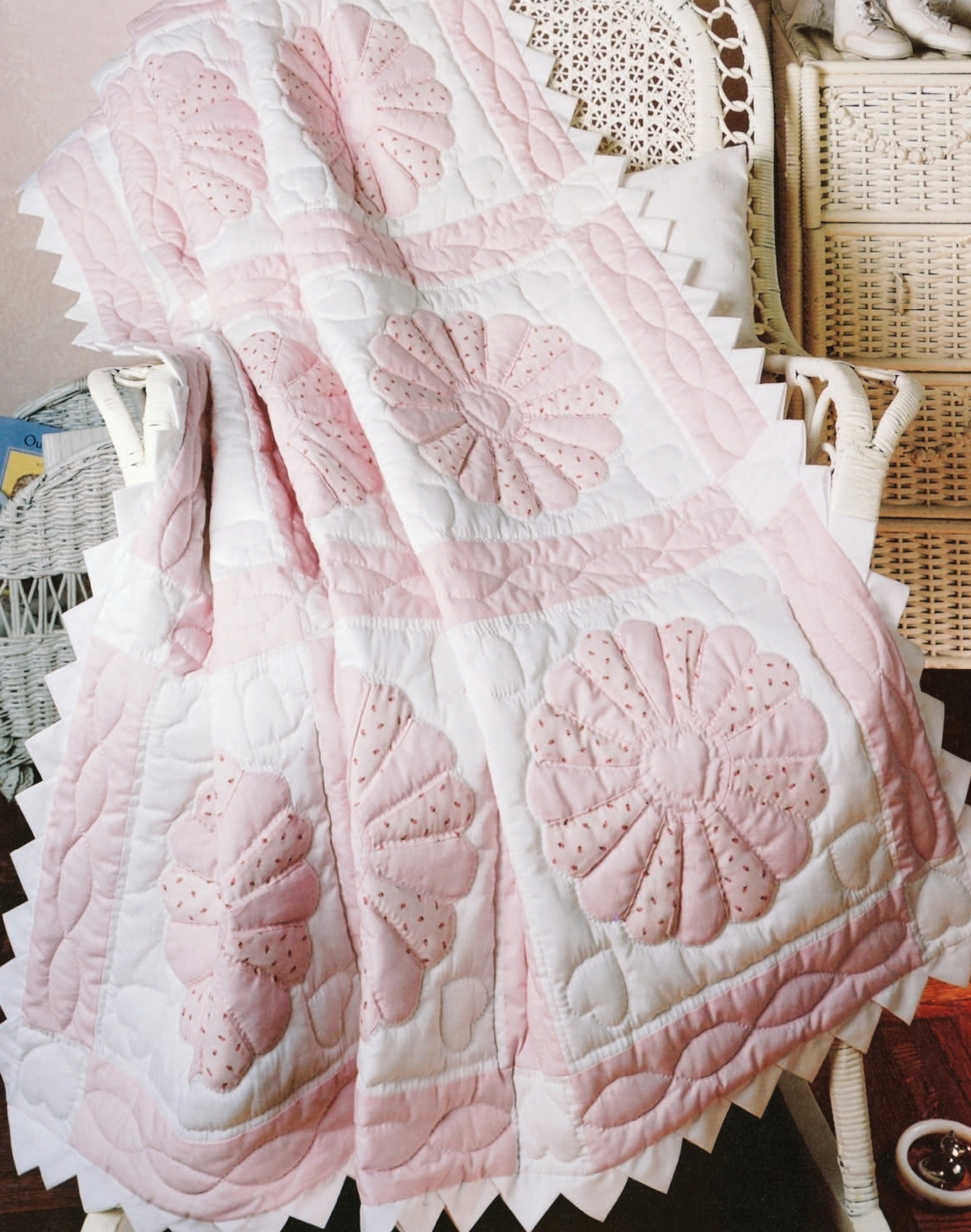 The Leisure Arts Rock-a-Bye Quilts, featuring pink and white flower patterns, is draped over a white wicker chair and surrounded by other wicker furniture in a cozy room—perfect for baby quilts.
