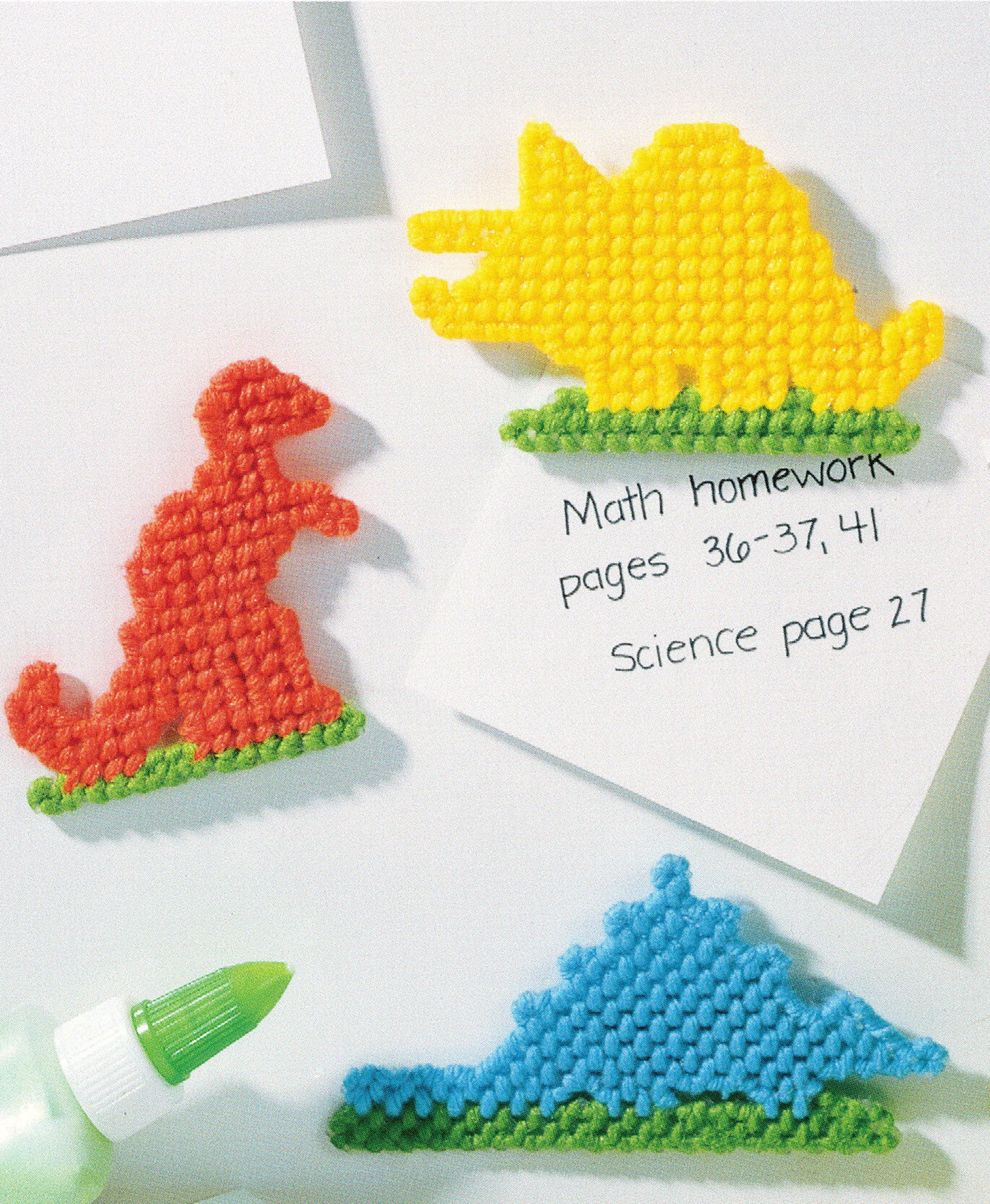 Three vibrant dinosaur memo holders from Leisure Arts’ “300+ Magnets in Plastic Canvas” are displayed on a white surface with a homework note, while a green glue tube adds extra color to these fun, magnetic creations.
