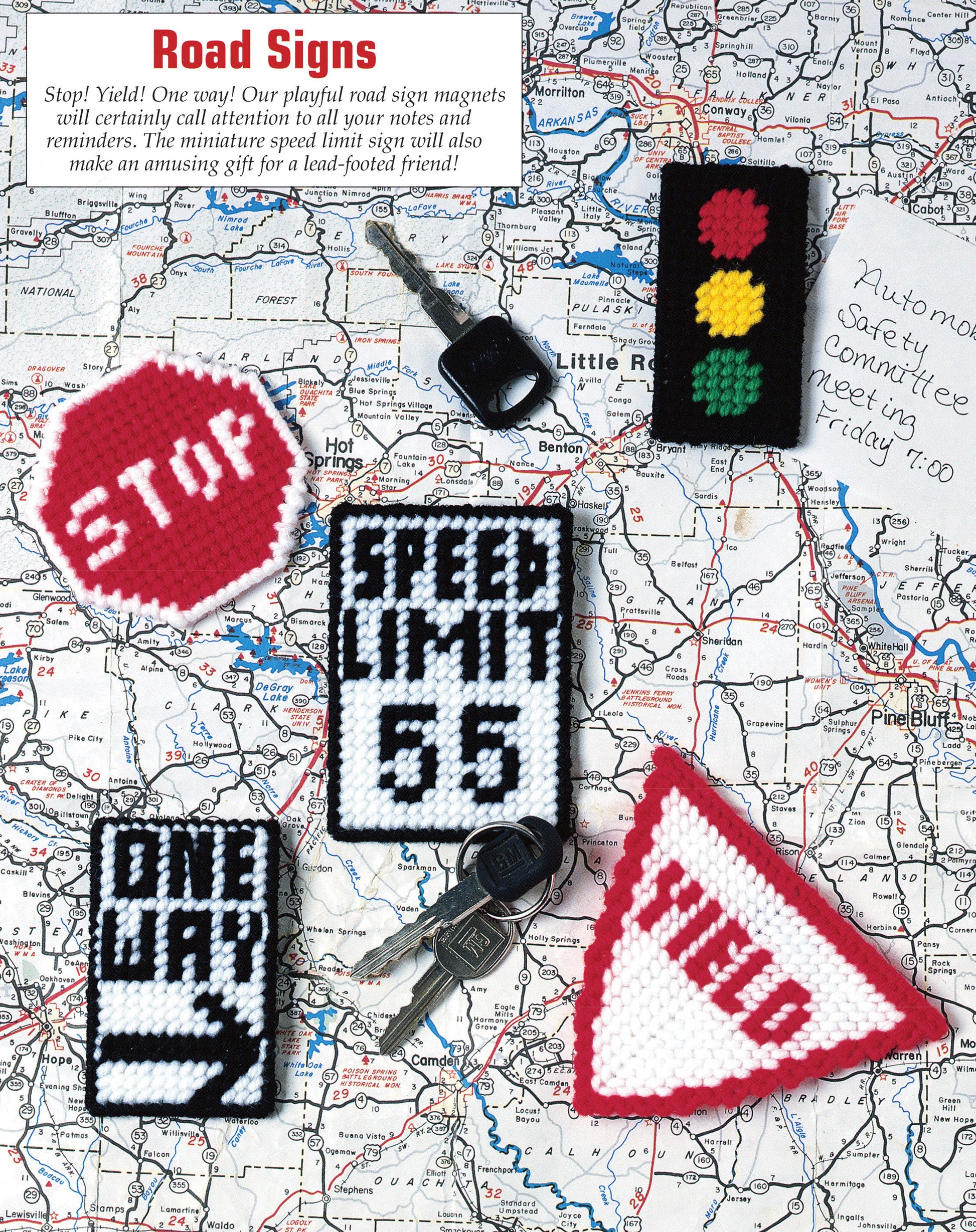 The image features keys, a handwritten note, and five handmade plastic canvas road sign magnets from Leisure Arts’ "300+ Magnets in Plastic Canvas," including stop, yield, speed limit 65, one way, and traffic light designs.
