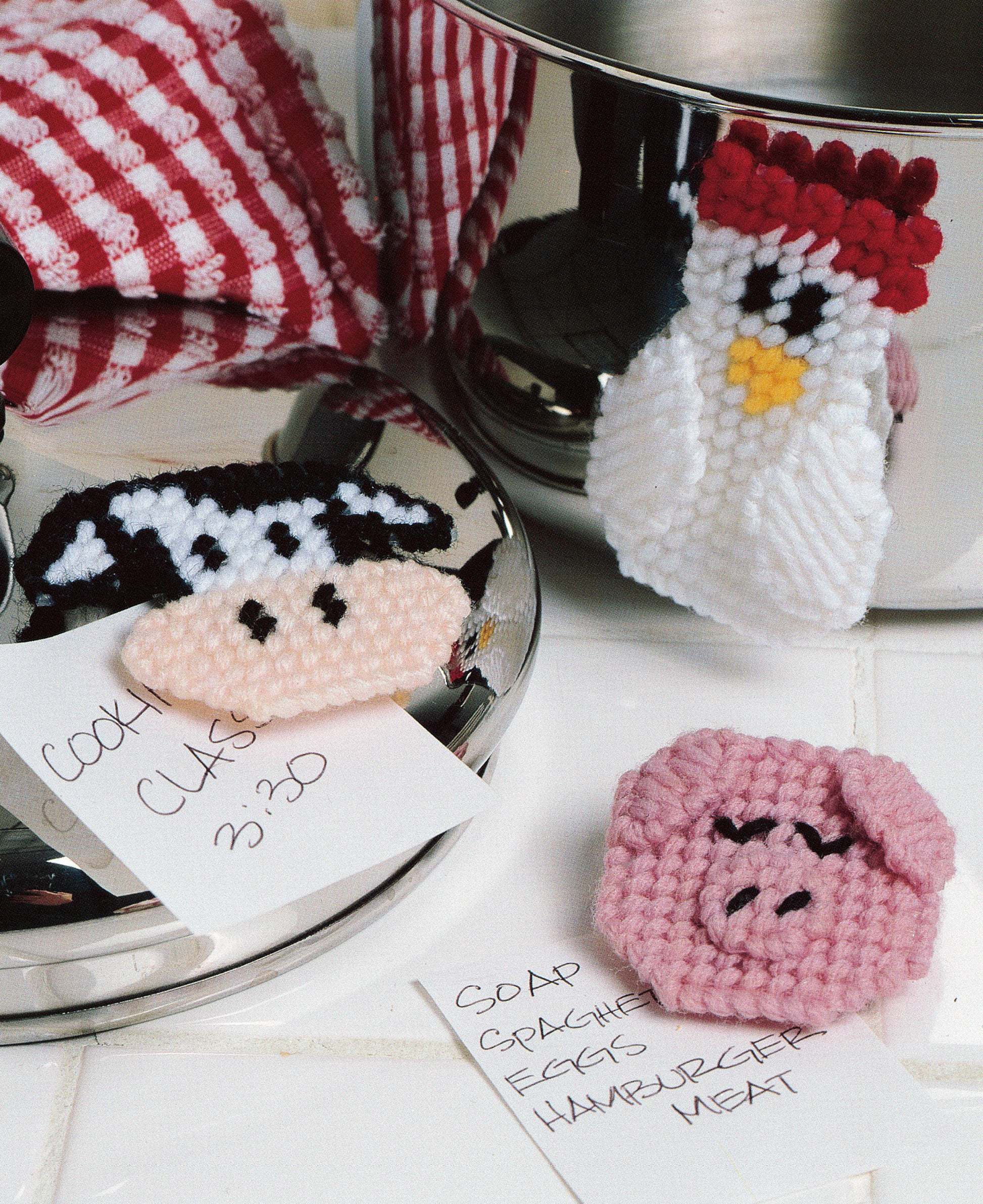 Three playful magnets from "300+ Magnets in Plastic Canvas" by Leisure Arts, shaped like a cow, chicken, and pig, rest on a counter near handwritten grocery notes and a red-checkered cloth. Their festive designs add charm to the kitchen scene.
