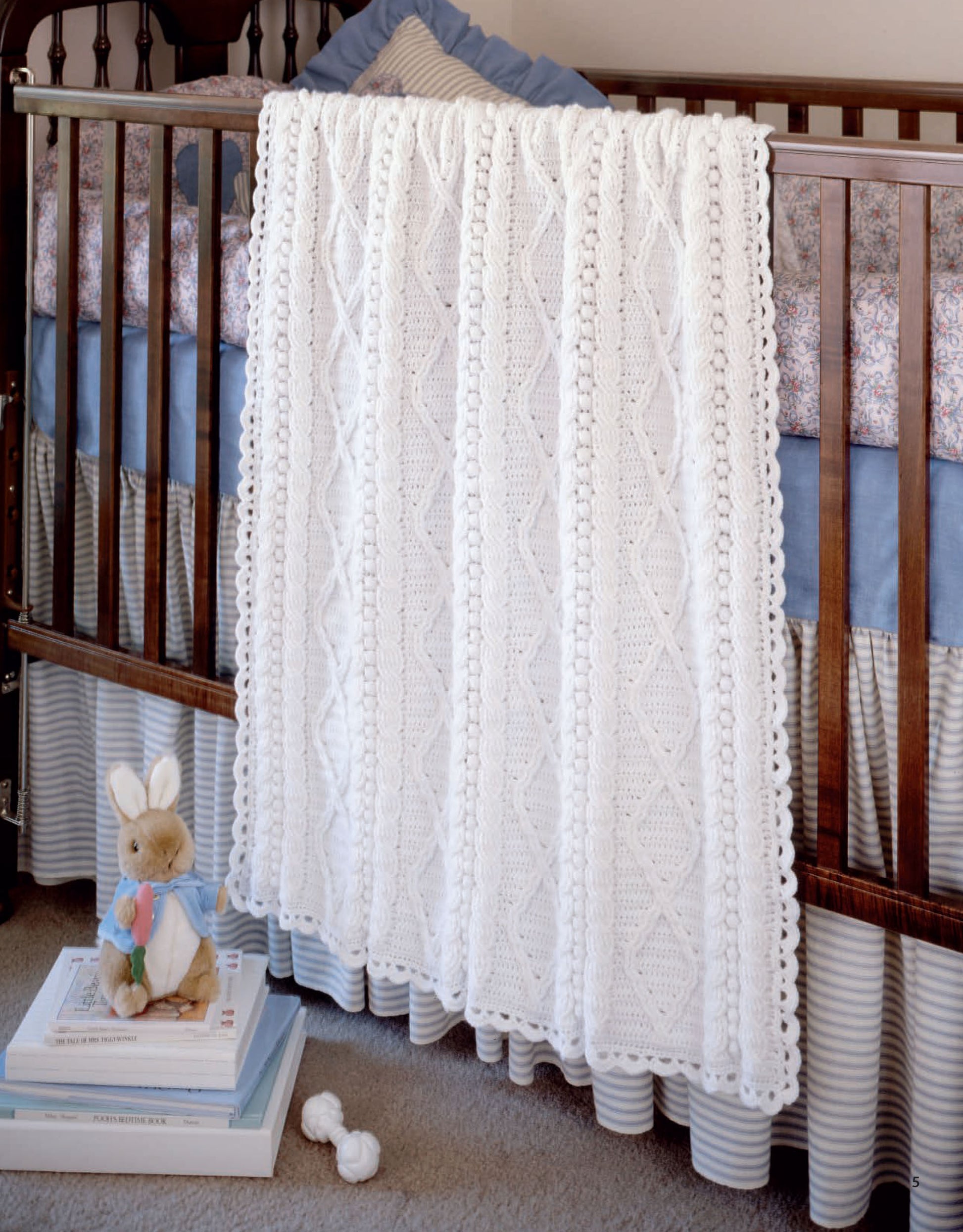A white, intricately knitted baby blanket from Leisure Arts’ "Aran Baby Afghans Digital Download" drapes over a wooden crib with a striped bed skirt, floral bedding, and a stuffed bunny placed on stacked books nearby.