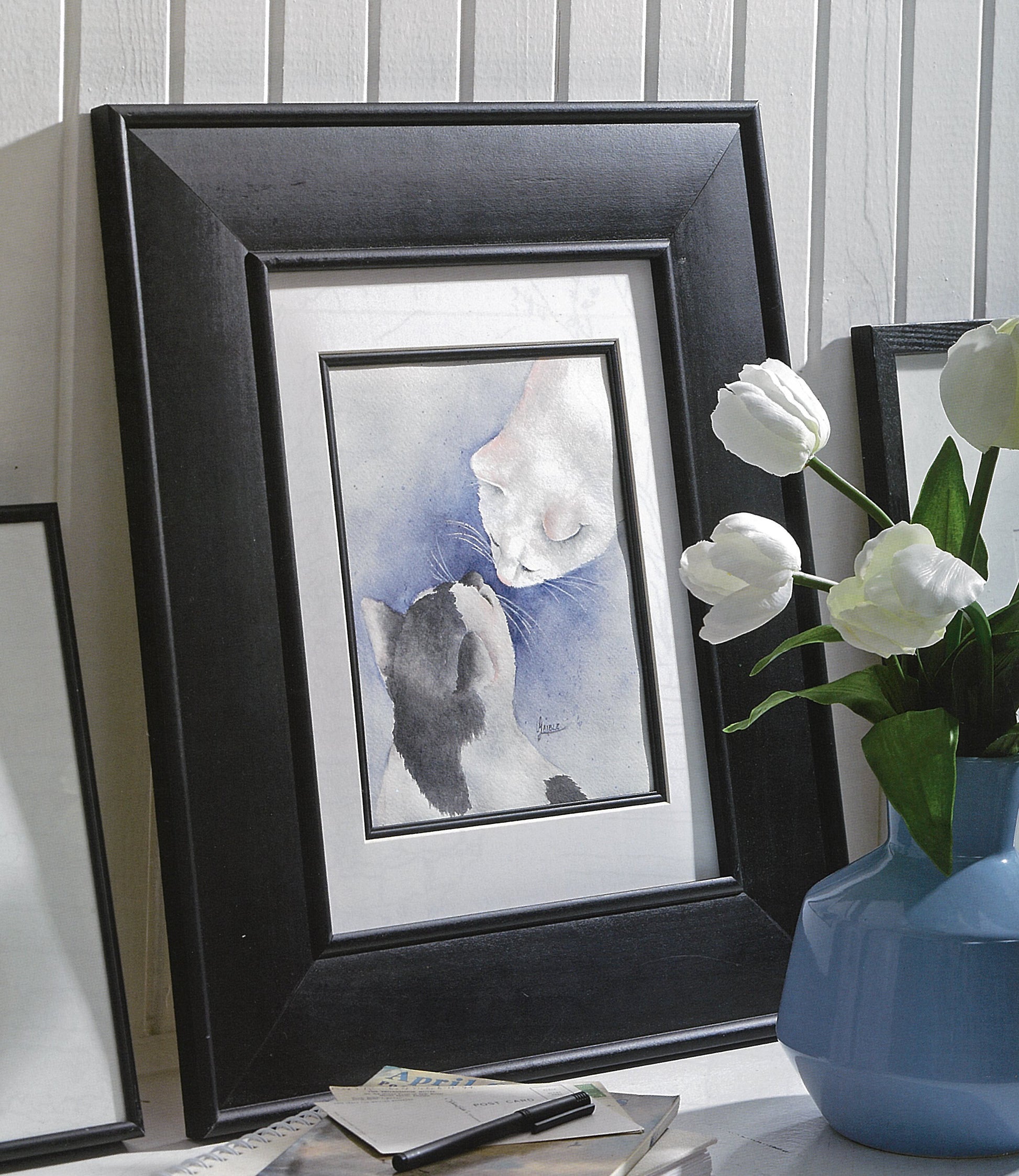 A framed painting of two cats touching noses, inspired by Watercolor Classics—7 Beginner Friendly Designs by Leisure Arts, sits on a shelf beside a blue vase with white tulips and some envelopes.