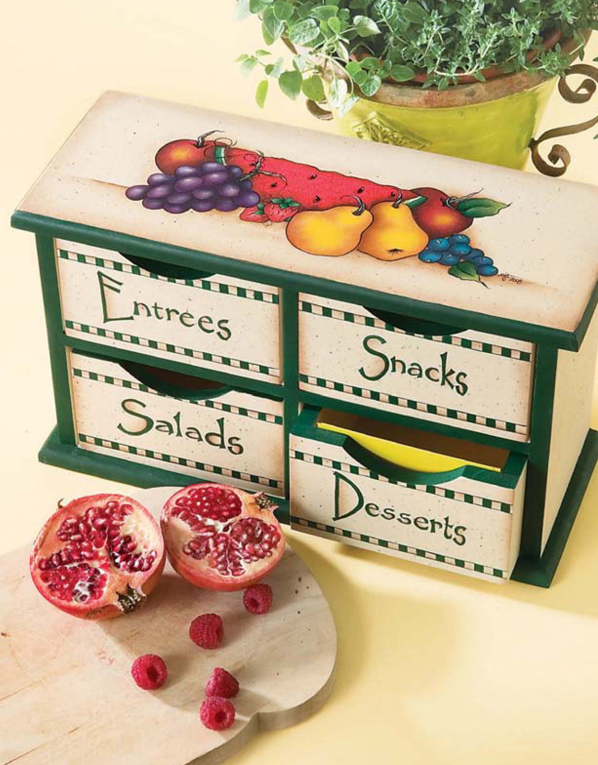 The Painted Kitchen by Leisure Arts is a small wooden drawer organizer labeled Entrées, Snacks, Salads, and Desserts with painted fruit accents, shown near a pomegranate, raspberries, and a green plant—ideal for charming DIY kitchen decor.
