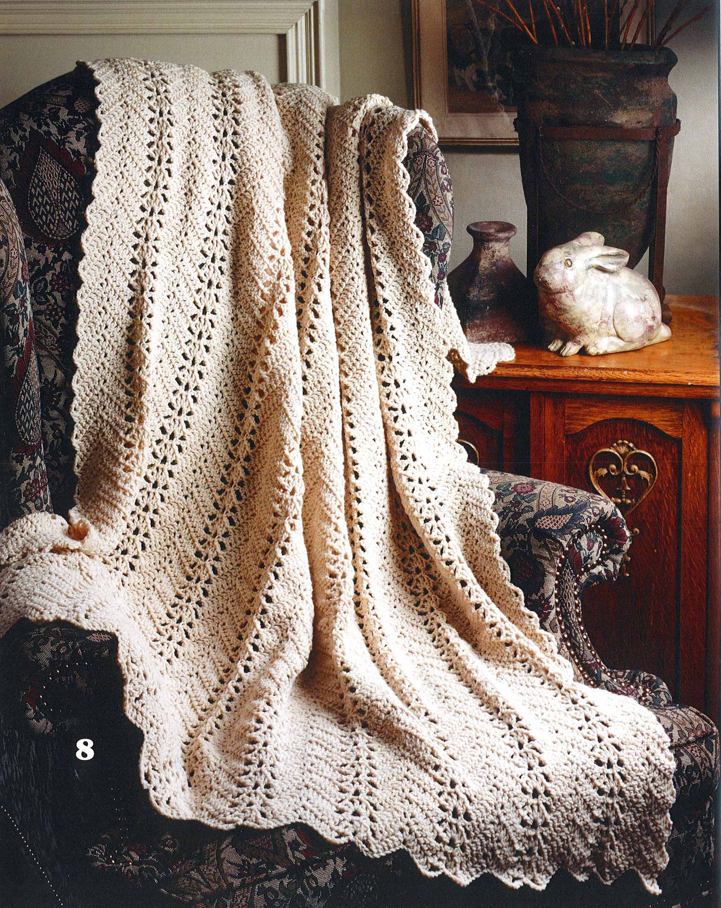 The Leisure Arts "Rippling Effects—16 Afghan Designs to Add to Your Home Decor" features cream, lacy crocheted afghans, perfect for draping over armchairs and adding elegance beside decor like vases and ceramic accents.