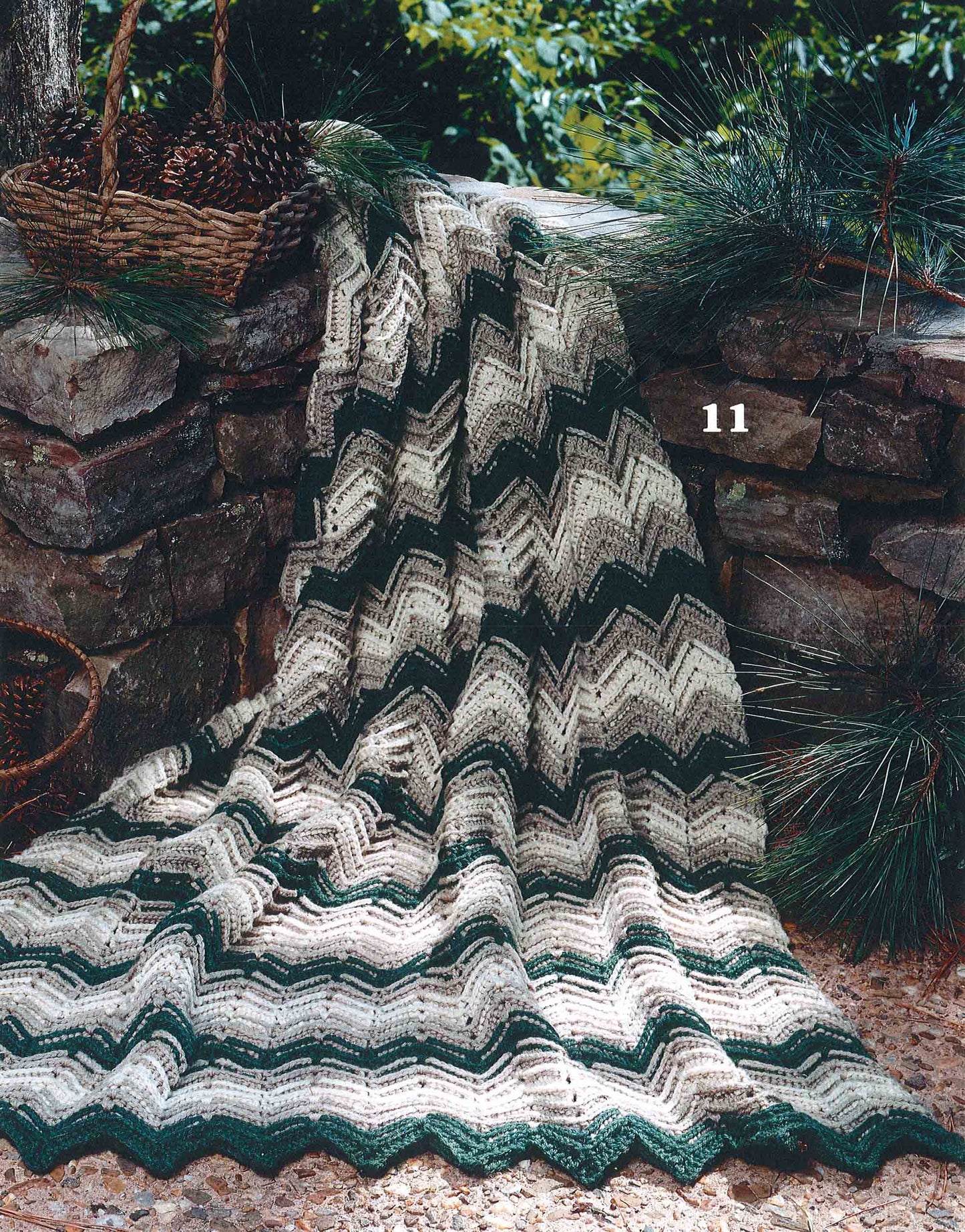 The Rippling Effects Afghan (#11) from Leisure Arts, crocheted in green, cream, and beige worsted yarn, drapes over a stone wall outdoors with pine branches, pine cones, and a wicker basket in the background.