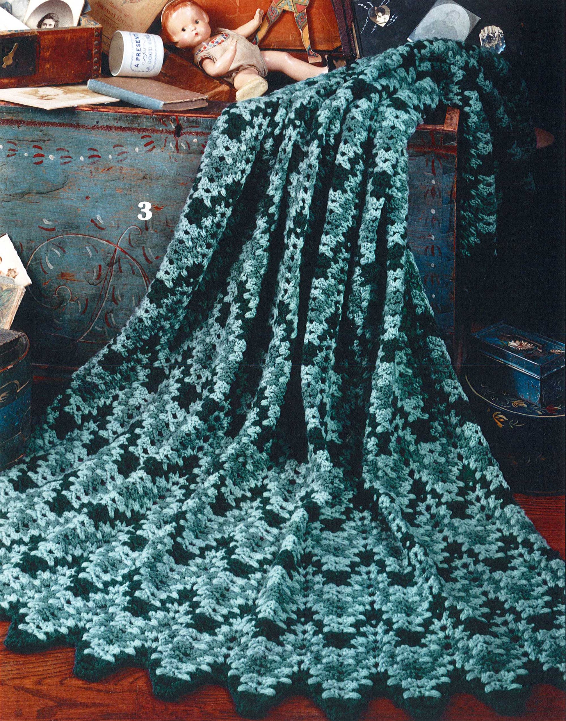 A large afghan, made with the Rippling Effects crochet pattern book by Leisure Arts, drapes over a blue trunk. Behind it are vintage items like a porcelain doll, tin, and cup.