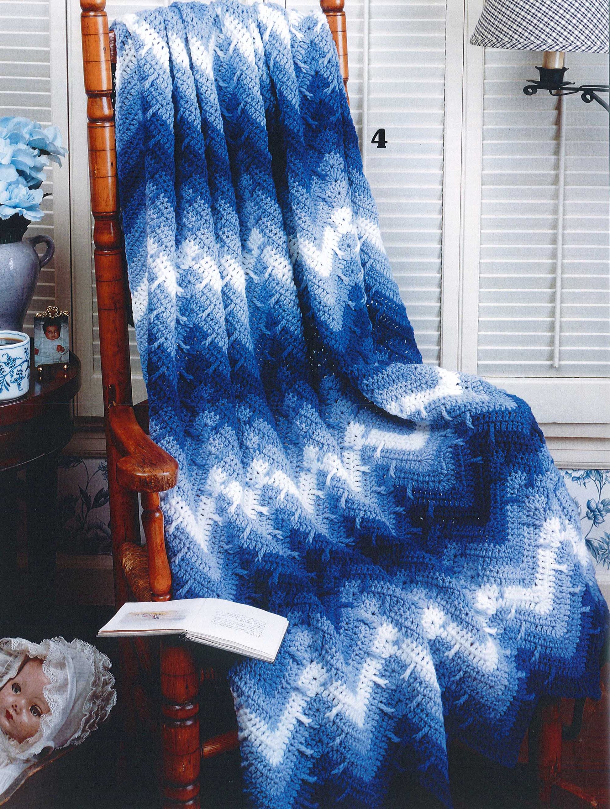 A wooden rocking chair draped with a blue and white zigzag afghan from "Rippling Effects—16 Afghan Designs to Add to Your Home Decor" by Leisure Arts sits next to a table with a teacup, flowers, an open book, and a doll on the floor.