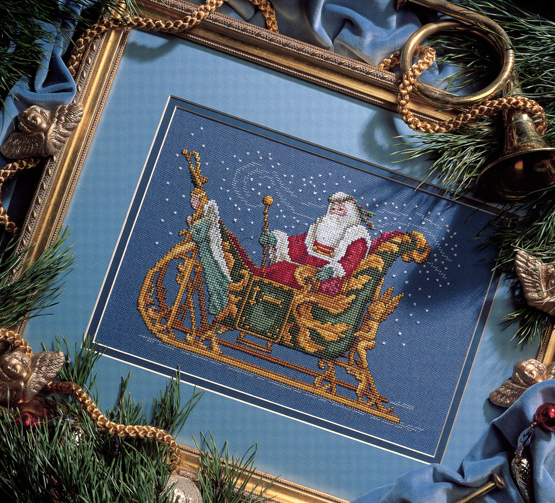 Celebrate the classic holiday tradition with Leisure Arts’ "Santa's Great Book of Cross Stitch," featuring festive patterns like Santa in a golden sleigh on blue fabric, surrounded by evergreens, gold ornaments, and snowflakes.