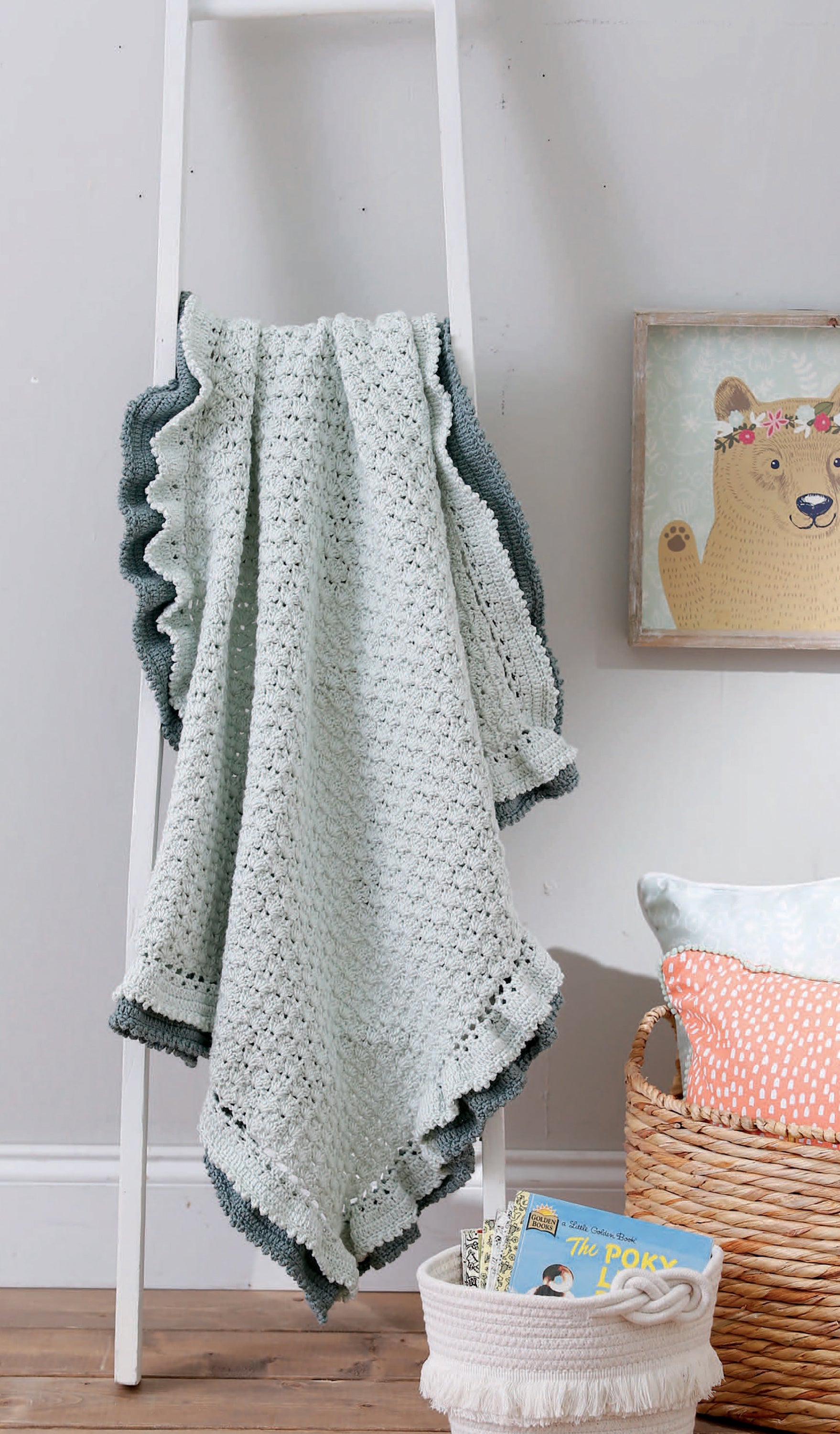 A light gray crochet baby blanket from Leisure Arts' "Our Best Baby Afghans" with a dark green border hangs over a white ladder. Beside it, baskets hold pillows, children's books, and a framed bear illustration on the wooden floor.