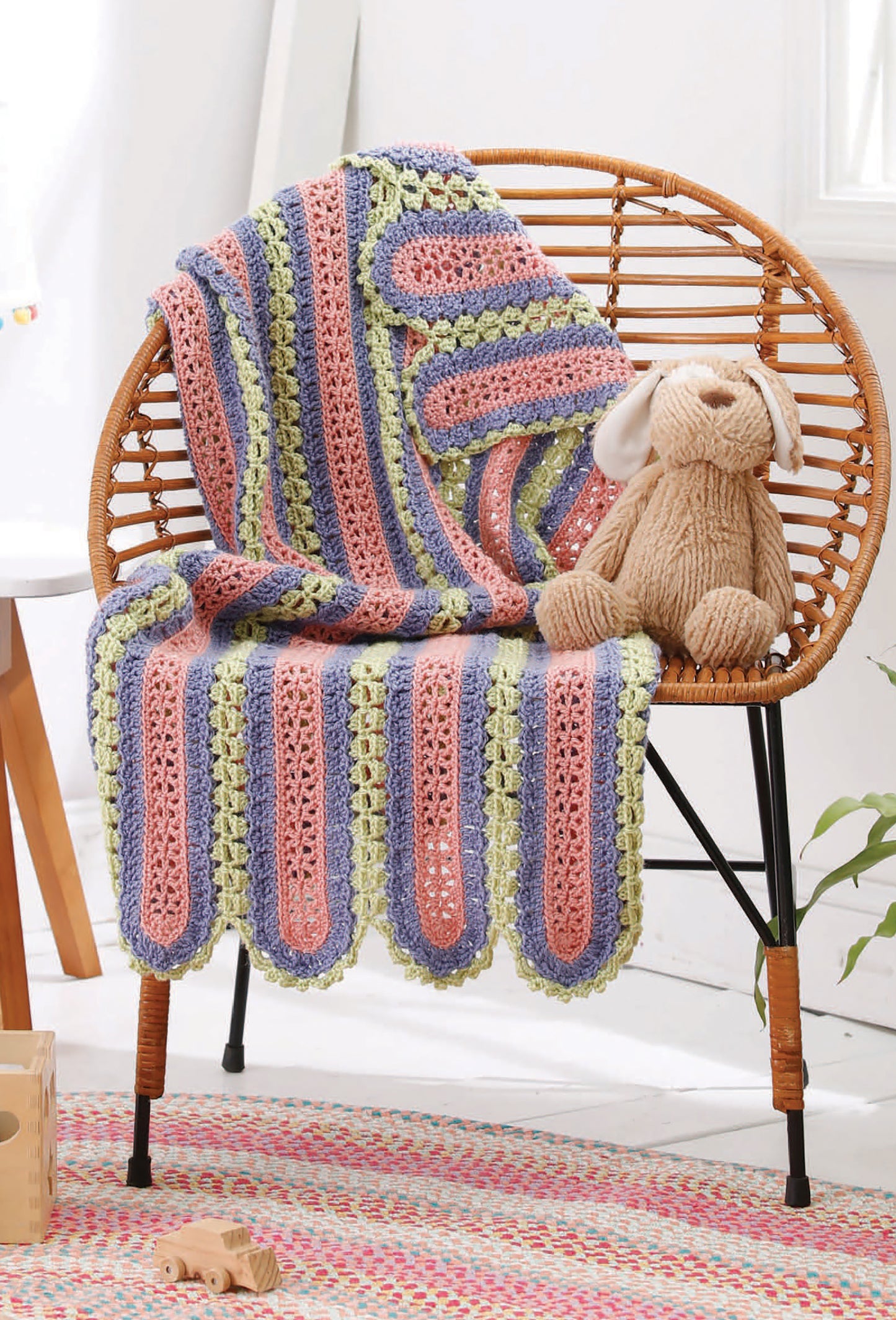 A striped baby blanket from Leisure Arts' "Our Best Baby Afghans," perfect for beginner crochet, drapes over a wicker chair next to a plush bunny, while wooden toys and bright light create a cozy room ambiance.