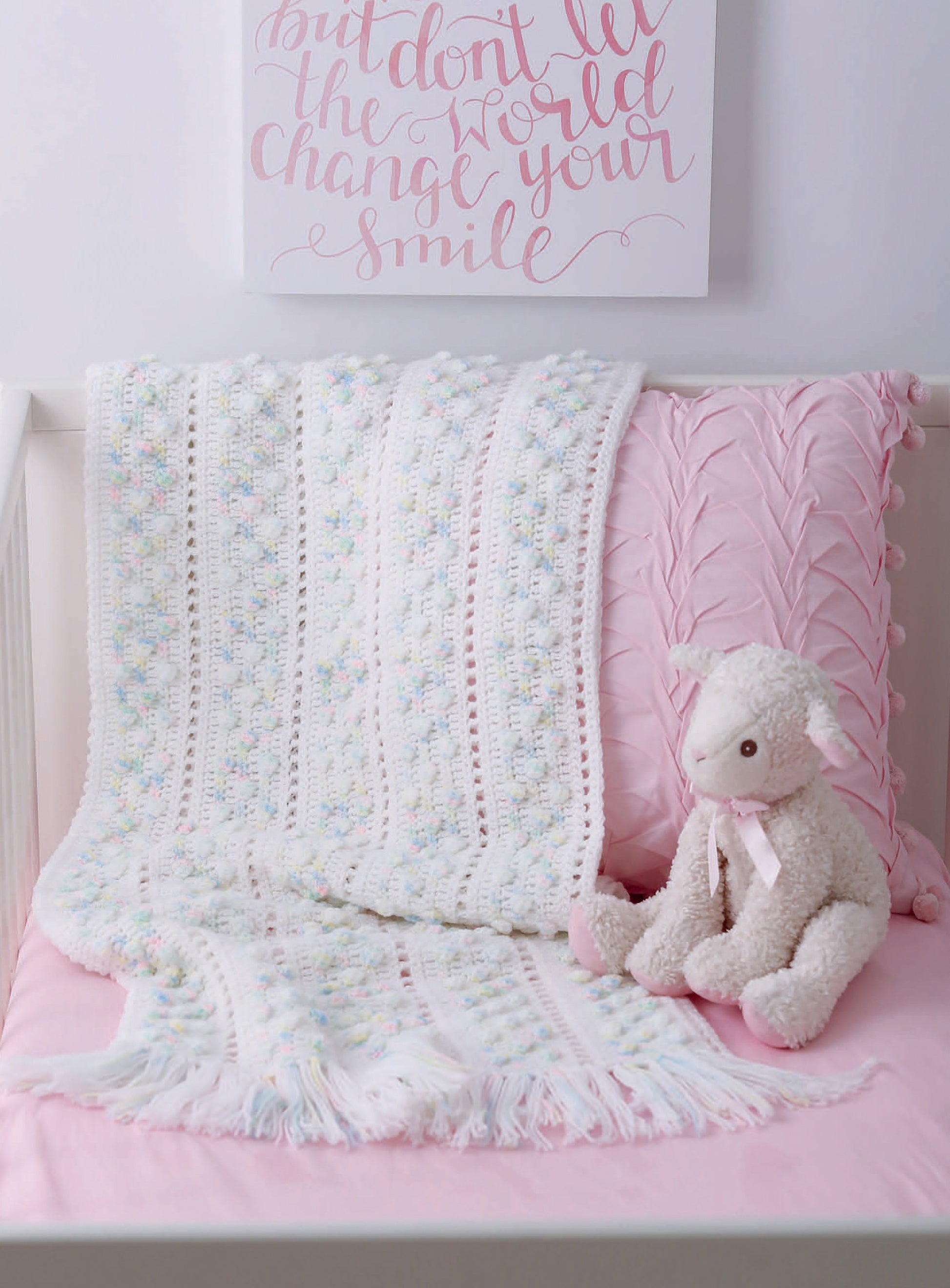 A white crib with a pink cushion, decorative pillow, and plush lamb toy features a soft baby blanket made from Leisure Arts' "Our Best Baby Afghans." Above the crib, a sign reads: But don’t let the world change your smile.