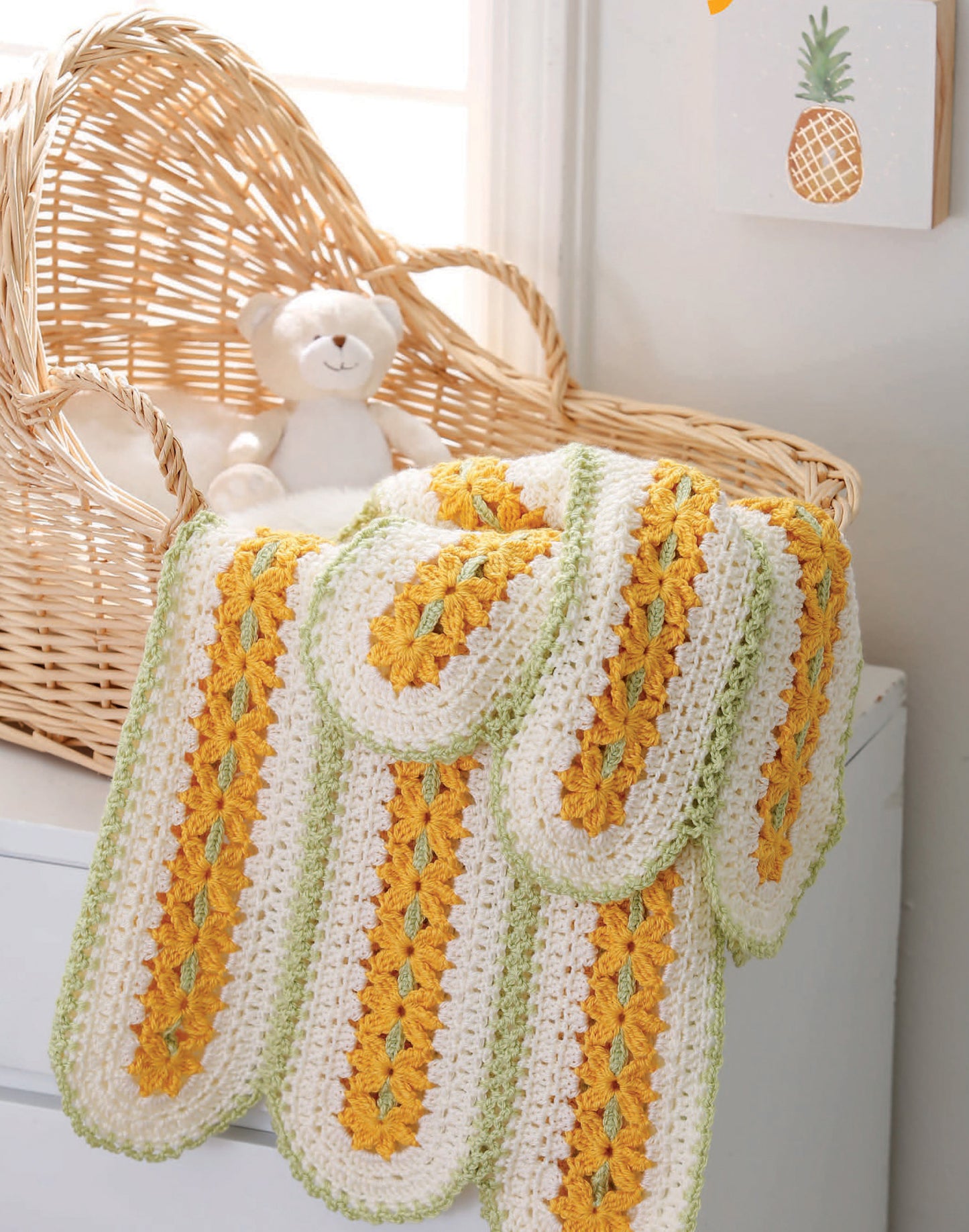 A cozy baby blanket from "Our Best Baby Afghans" by Leisure Arts with white, yellow, and green stripes is draped over a wicker basket with a plush bear, set by a sunny window—perfect for beginner crochet enthusiasts.