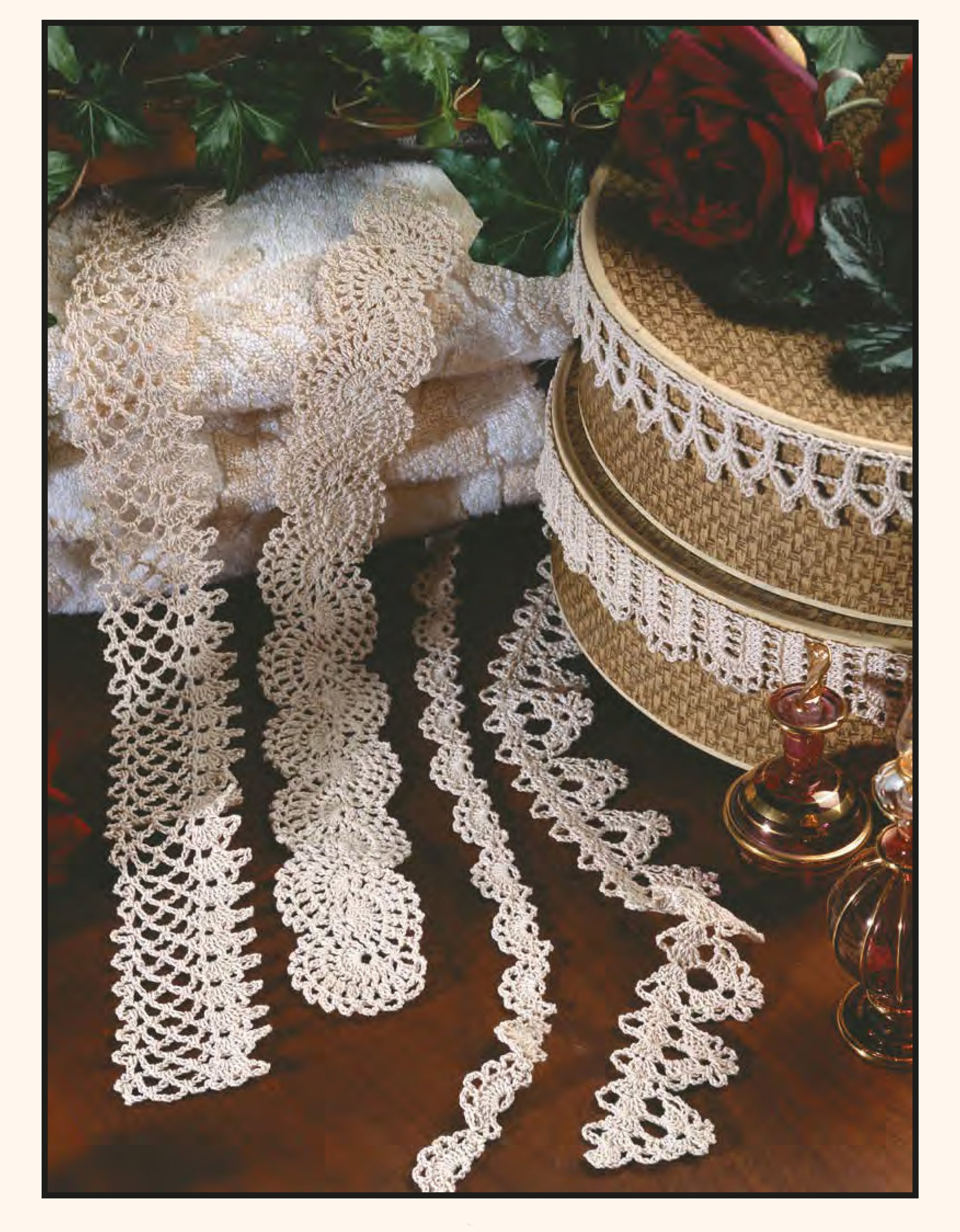 Four cream lace strips from Leisure Arts' 111 Easy Edgings crochet patterns are arranged on a brown surface with two lace-trimmed, rose-adorned boxes and small glass bottles nearby. Towels and greenery appear in the background.