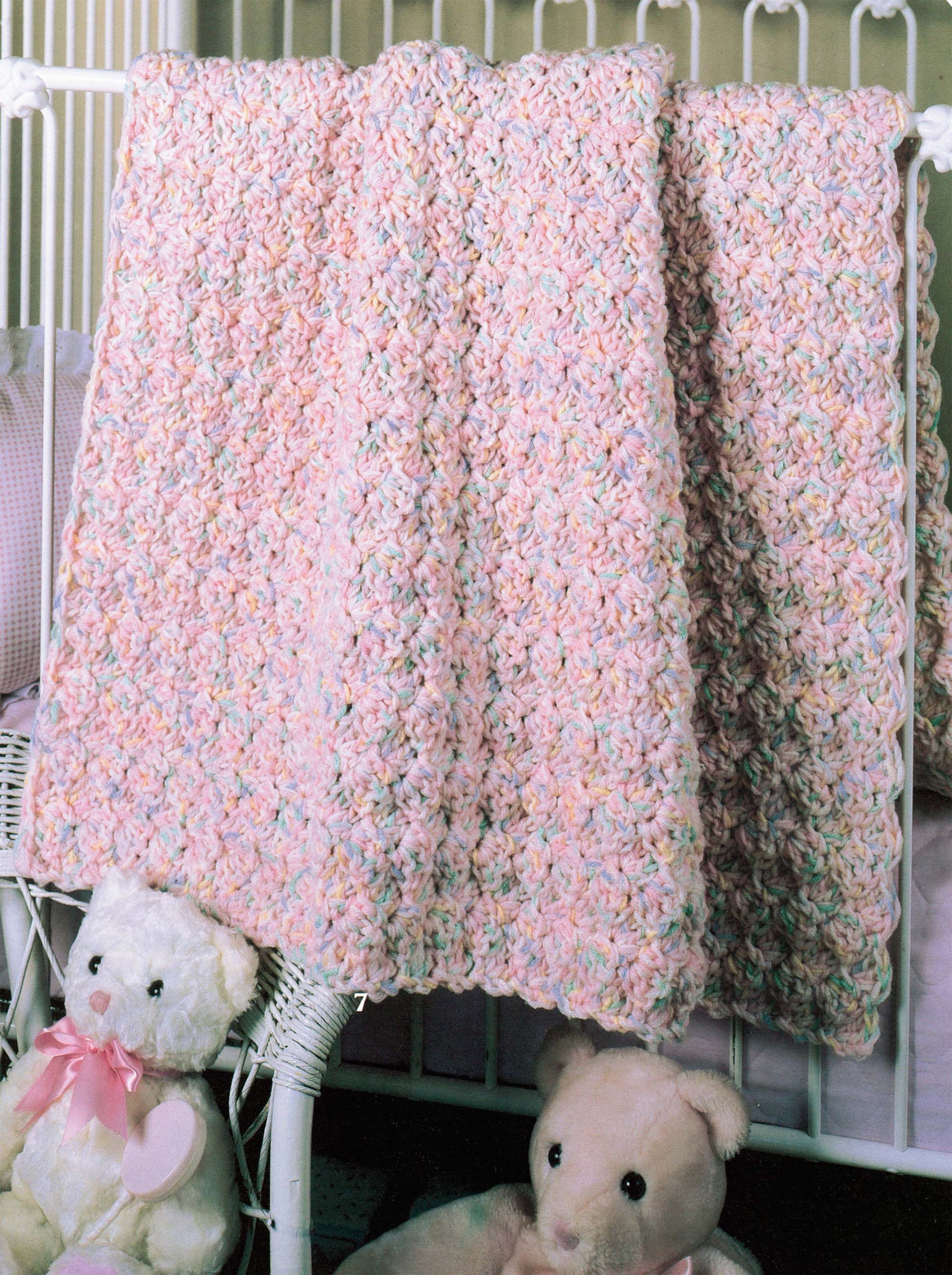 The "More Double-Quick for Baby" crochet blanket by Leisure Arts, in soft pastels, hangs on a white crib, with two plush teddy bears resting on a wicker chair nearby.