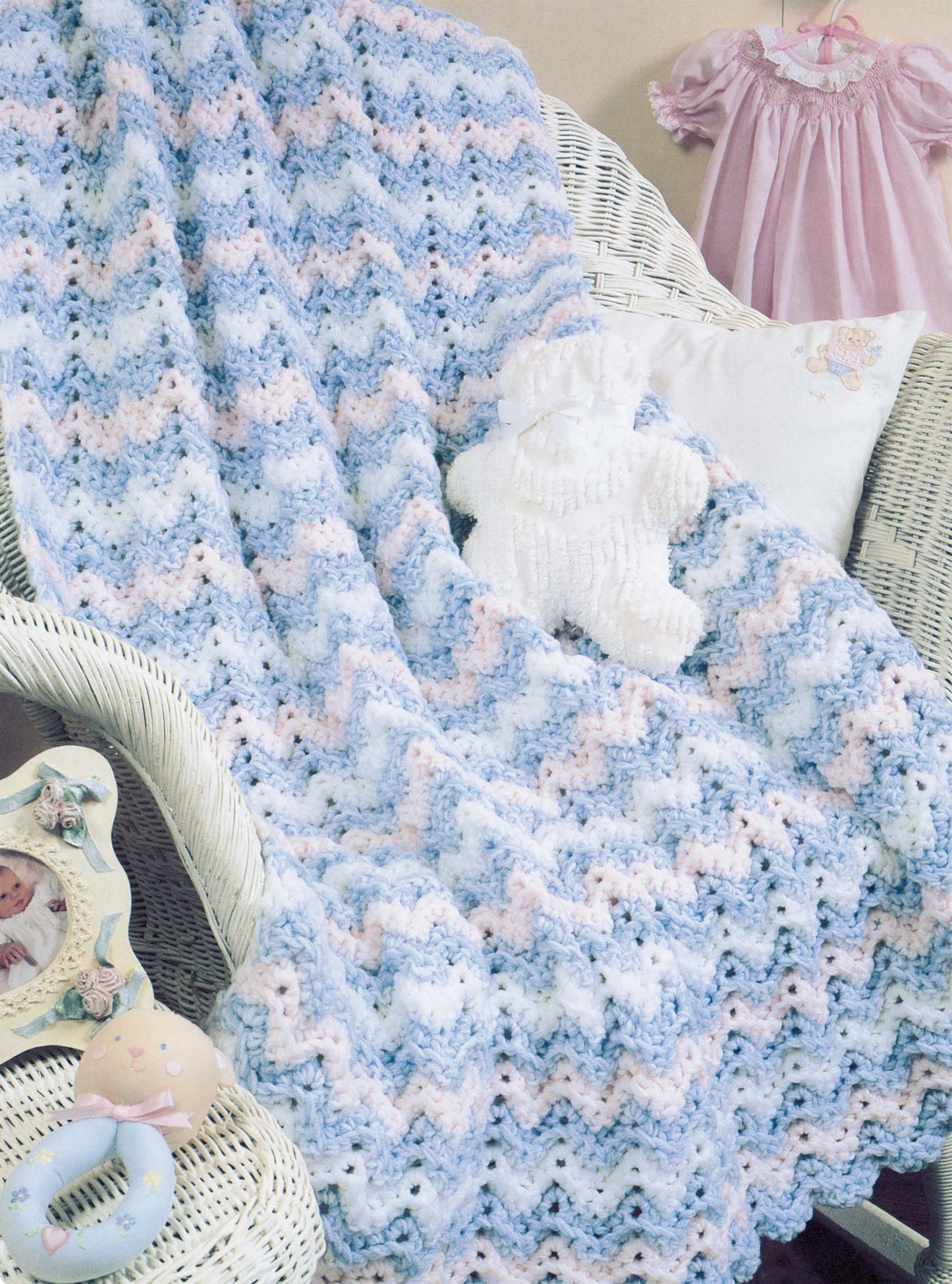 A pastel chevron crochet blanket from Leisure Arts’ "More Double-Quick for Baby" is draped over a white wicker chair with nursery decor—ideal for beginner crochet enthusiasts looking for new baby blanket patterns.