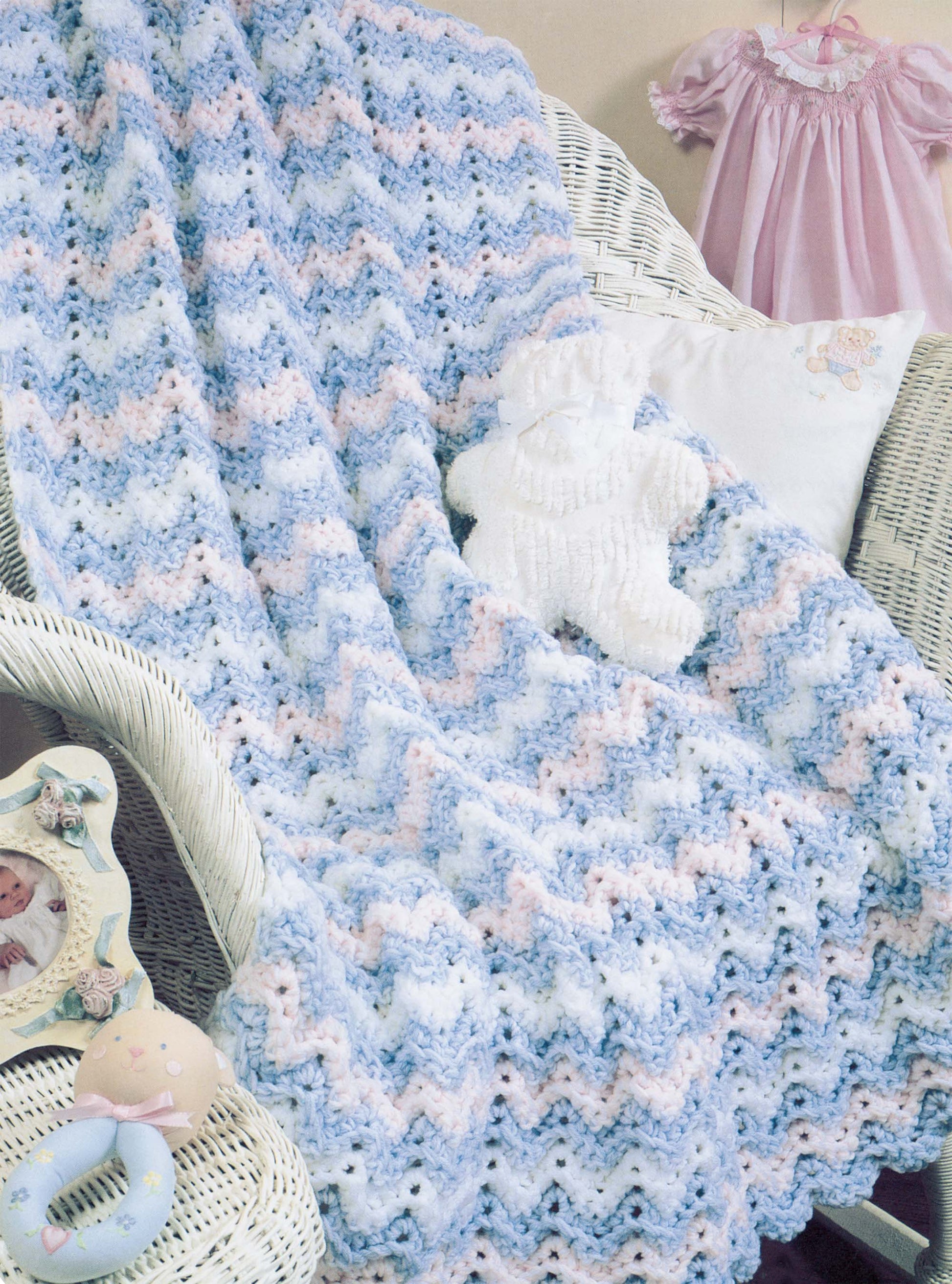 A pastel chevron crochet blanket from Leisure Arts’ "More Double-Quick for Baby" is draped over a white wicker chair with nursery decor—ideal for beginner crochet enthusiasts looking for new baby blanket patterns.
