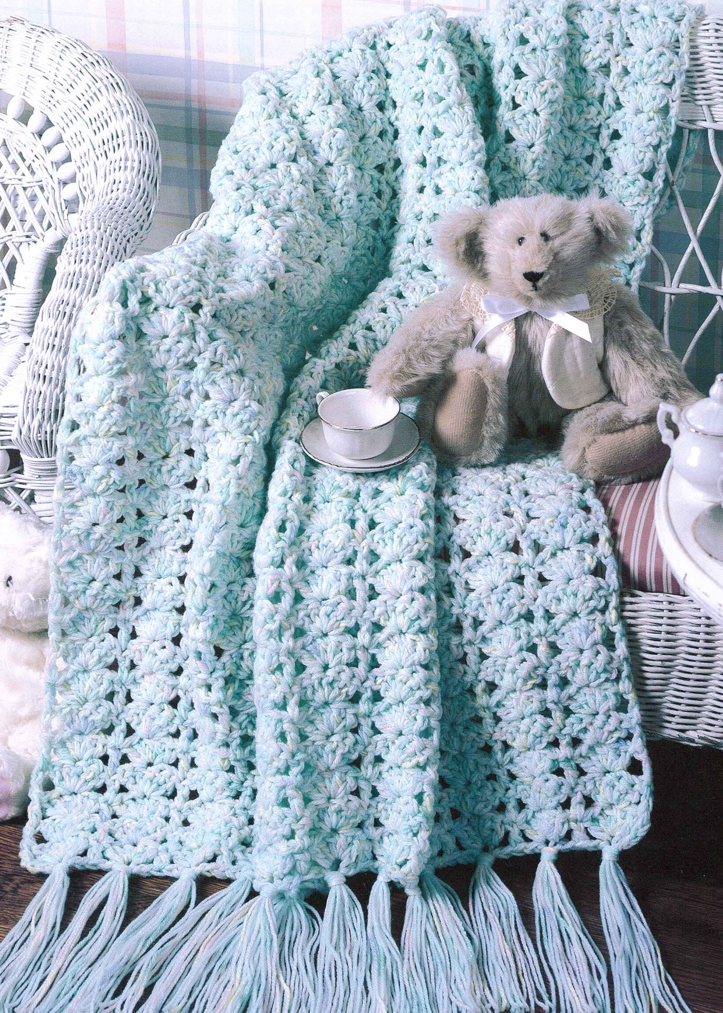 A pale blue crocheted blanket from More Double-Quick for Baby by Leisure Arts drapes over a white wicker chair. A plush teddy bear with a ribbon and a teacup rest on the soft fringe, with a teapot nearby.