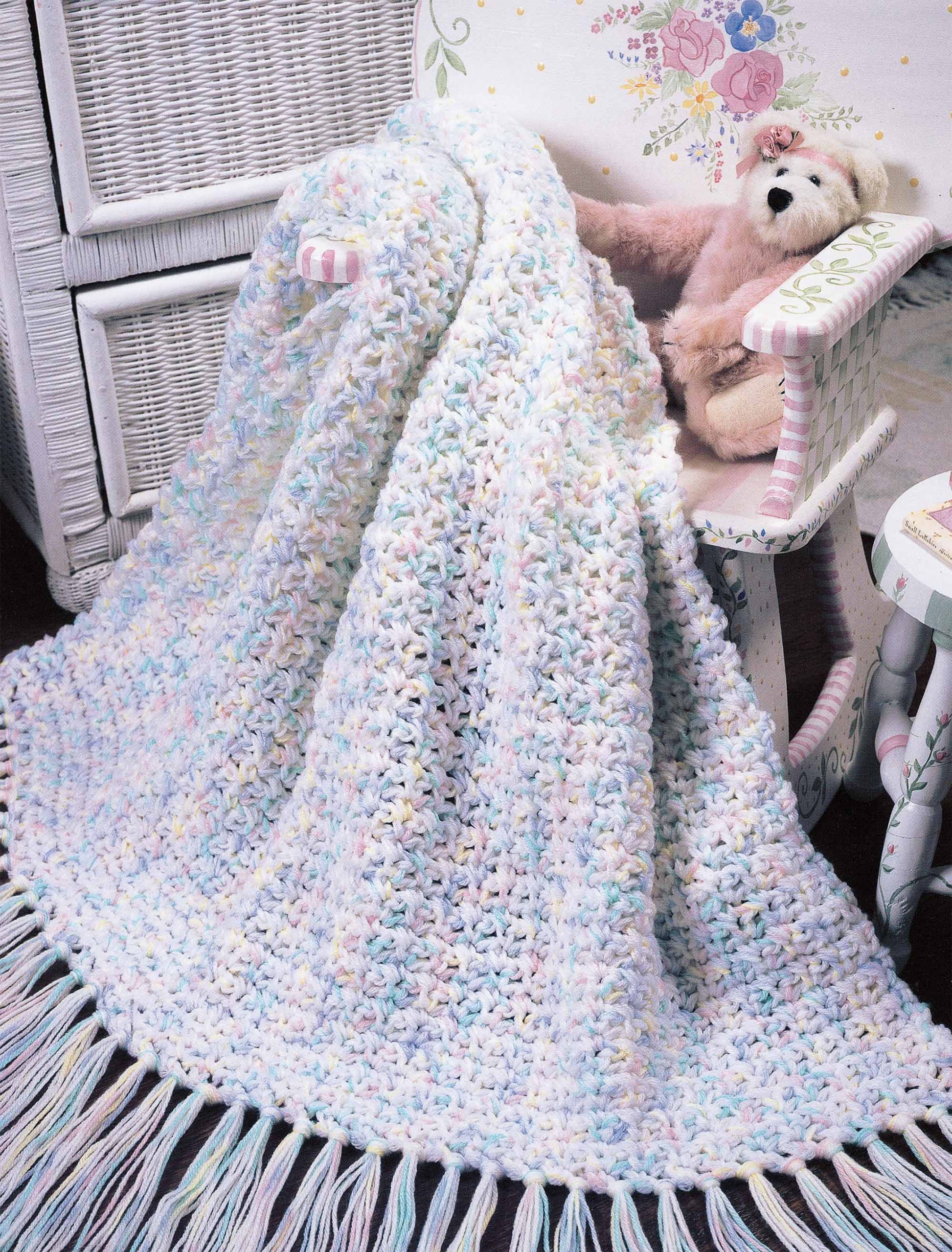 A pastel crocheted blanket made with worsted weight yarn from Leisure Arts' "More Double-Quick for Baby" drapes over a white wicker chair, while a rocking chair holding a teddy bear sits in a nursery with floral wall decorations.