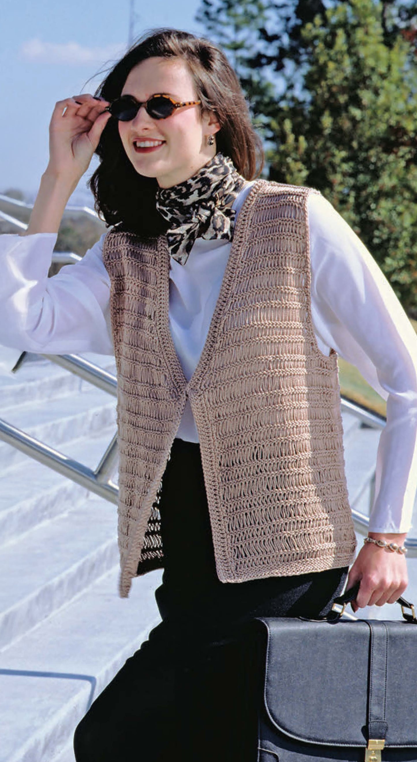 A woman wearing sunglasses, a patterned scarf, a white blouse, and a beige crochet vest inspired by Leisure Arts’ The Big Book of Quick Knit Sweaters smiles outdoors while holding a black bag.