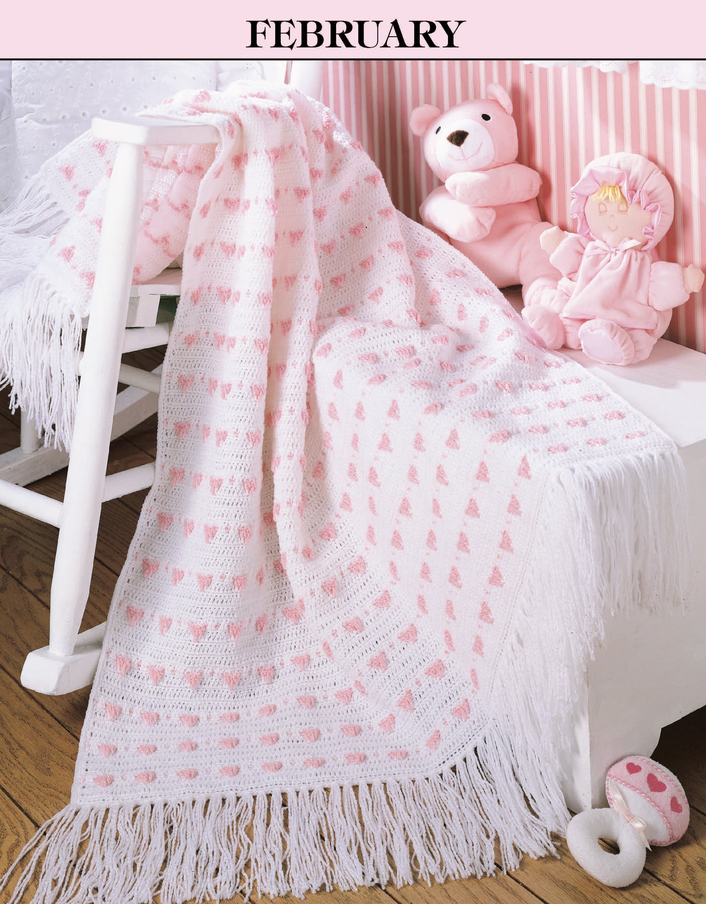 A white fringed baby blanket with pink heart motifs, from "A Year of Baby Afghans, Book 2" by Leisure Arts, drapes over a rocking chair in a pink-accented nursery with teddy bears and a doll. "February" is displayed above.