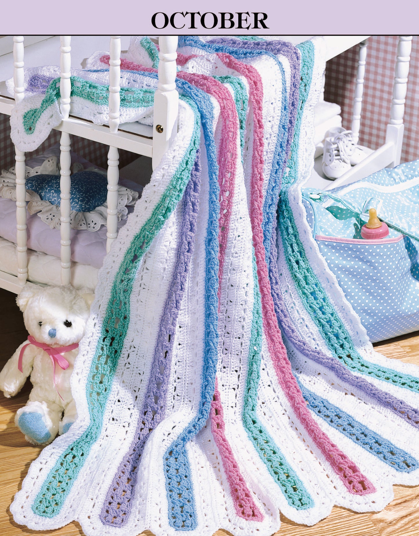 A cozy blanket from "A Year of Baby Afghans, Book 2" by Leisure Arts with pastel stripes is draped over a white crib. A teddy bear with a pink bow sits beside a blue polka-dot bag, highlighting the "October" themed baby afghan scene.