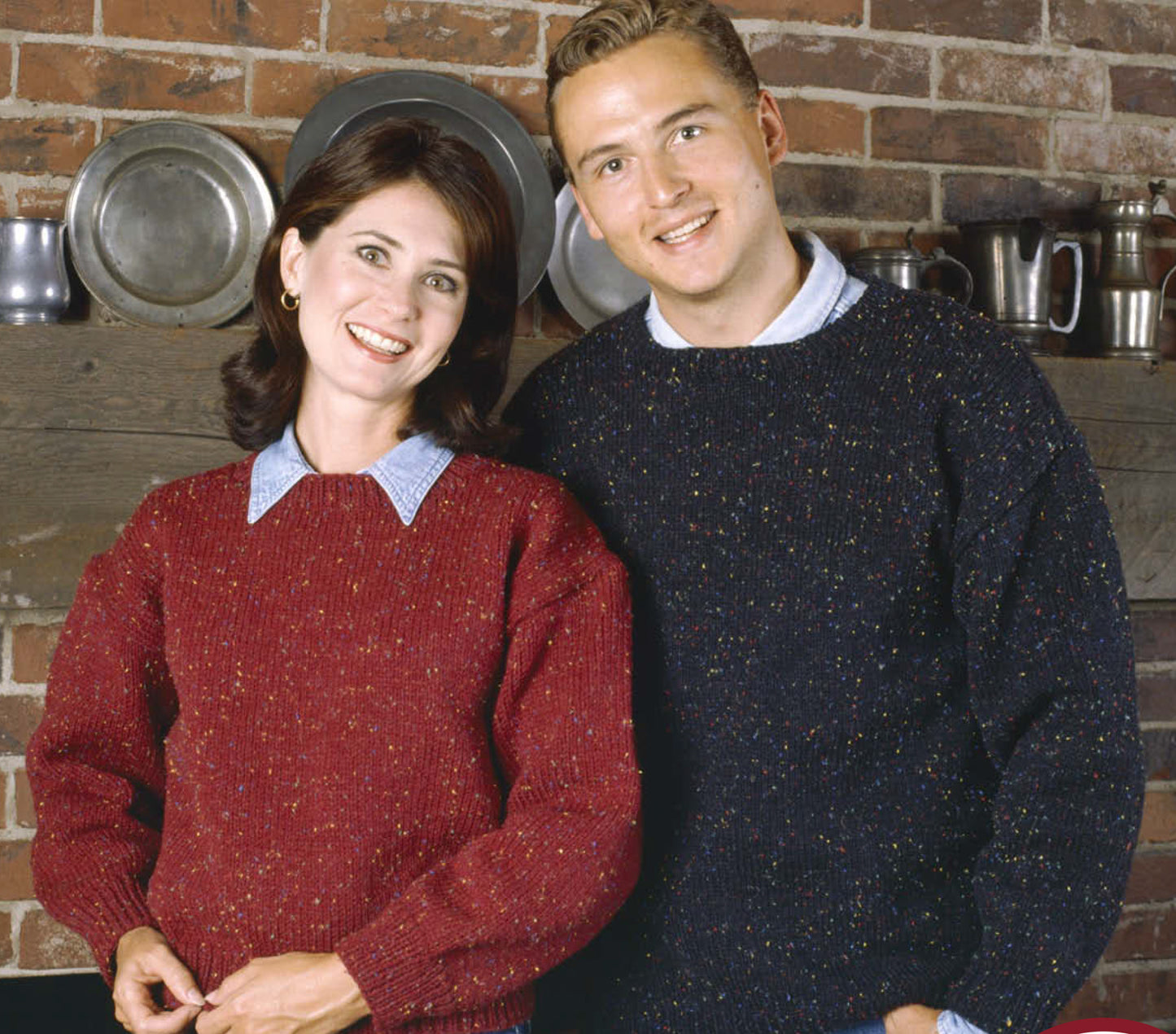 A woman in a red sweater from the Our Best Knit Collection by Leisure Arts stands smiling with a man in a dark sweater, both in front of shelves lined with metal pots and plates against a brick wall.