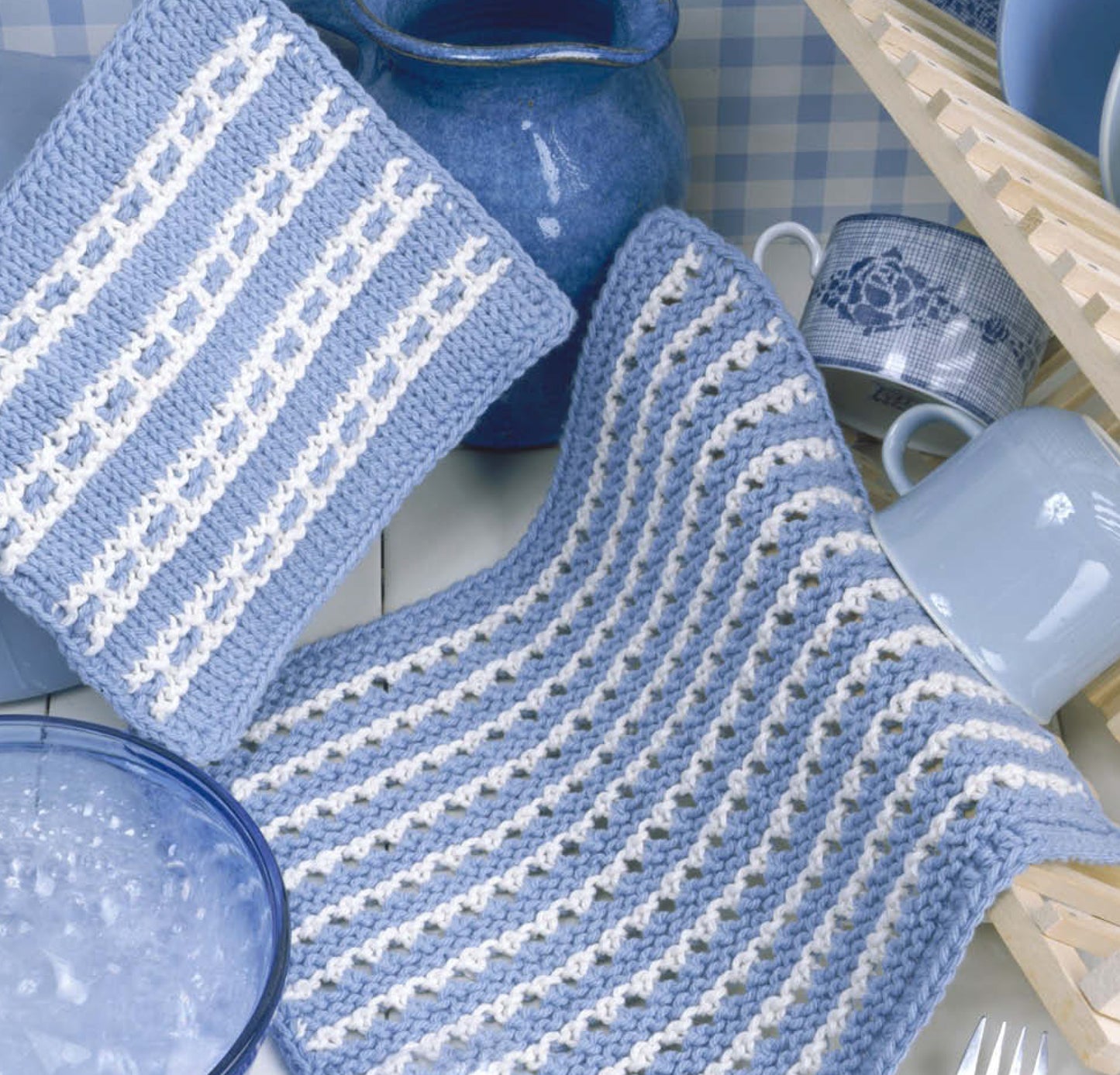 The "Our Best Knit Collection" by Leisure Arts features two blue and white knitted dishcloths, ideal for beginner or easy projects, displayed with blue dishes, a floral teacup, and a drying rack on a blue checked background.