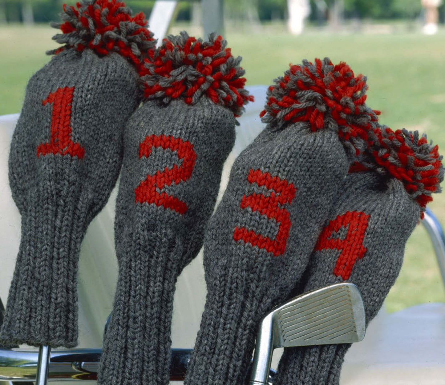 Four gray knit golf club headcovers with red numbers and red-gray pom-poms top clubs on a cart against a green field—an ideal showcase for Leisure Arts' "Our Best Knit Collection" easy or beginner knitting patterns.
