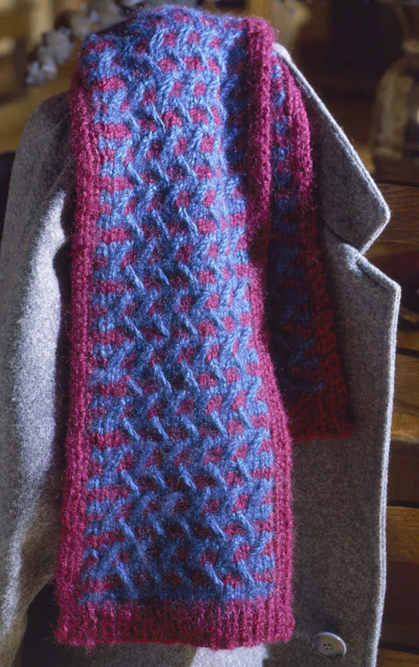 The Leisure Arts Our Best Knit Collection features a geometric blue and burgundy scarf with burgundy edges, draped over a gray wool coat—an ideal, easy project for beginner knitters.