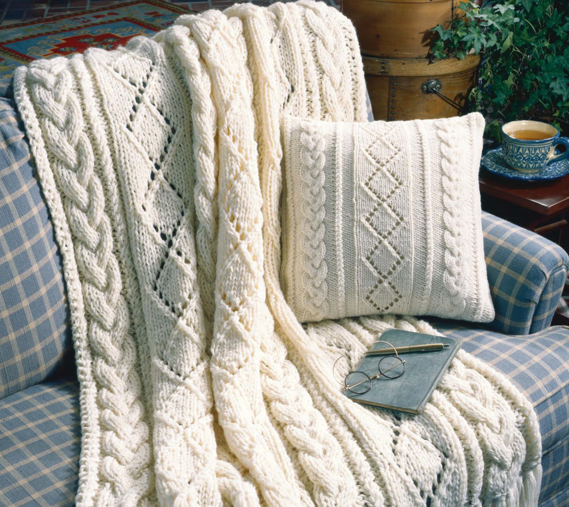 The "Our Best Knit Collection" by Leisure Arts features a cream knitted blanket and matching pillow with cable and diamond patterns, draped on a blue plaid sofa beside a book, glasses, and a cup of tea on a wooden table.