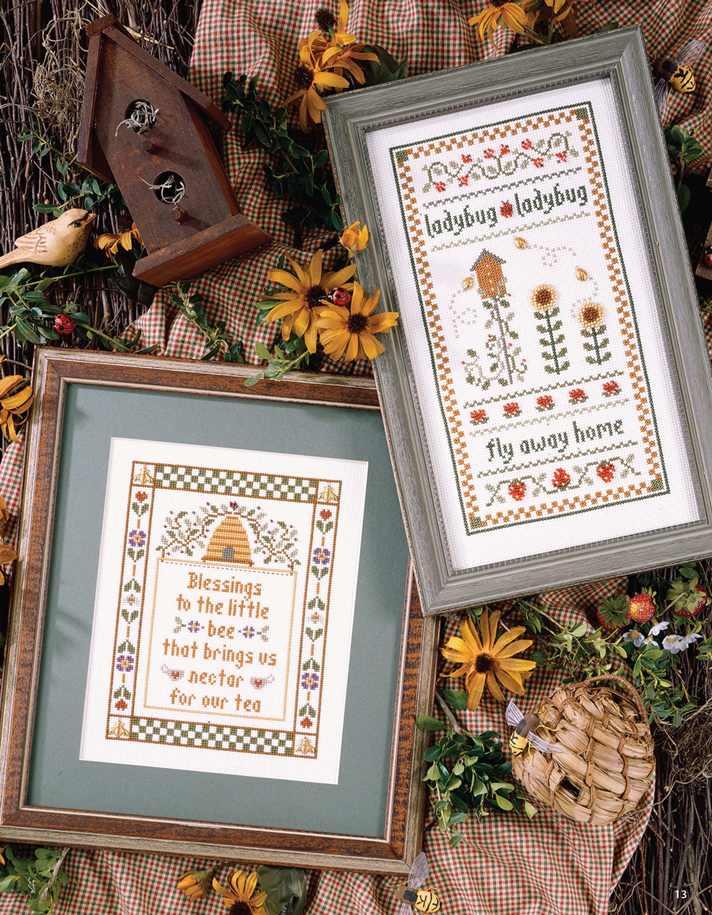Two framed cross-stitch pieces made with Leisure Arts' Joy in the Journey charts rest on a checked cloth amid faux flowers, a birdhouse, and woven bee skep—one features bees and a hive; the other shows ladybugs, blooms, and “Fly away home.”.